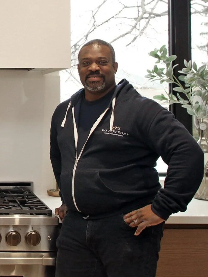 Chris is standing in a kitchen near a stove, wearing a black hoodie with 'Waterfront' logo, smiling, with a window and plant behind him.