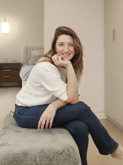 Waterfront Home Improvements owner Julie is sitting on a bench, smiling and resting her chin on her hand, with a lamp and nightstand in the background.