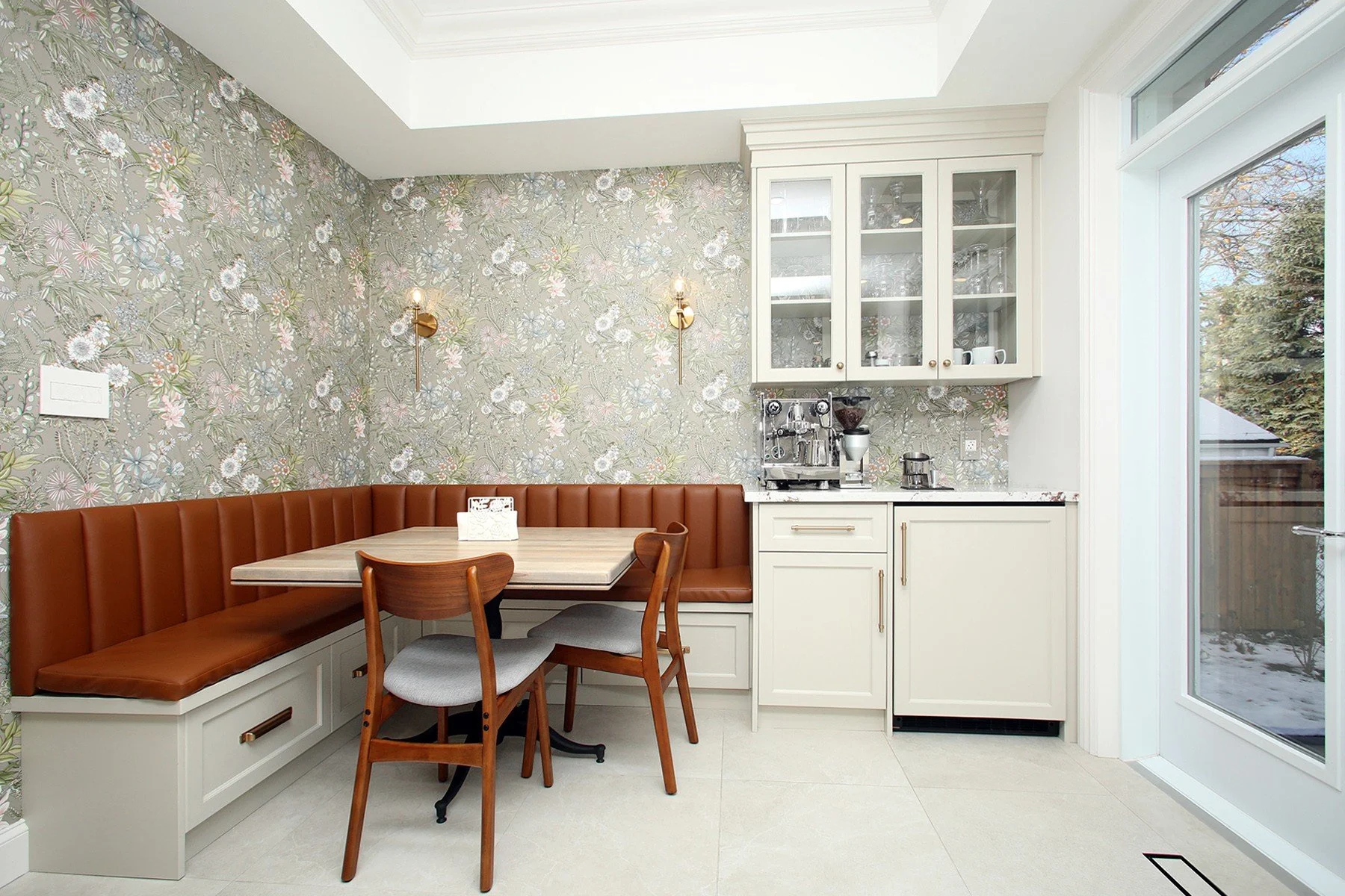 Kitchen nook with floral wallpaper, U-shaped beige leather bench, wooden table, two wooden chairs with upholstered seats, white cabinets, coffee machine, coffee grinder, large window showing outdoor trees and snow.