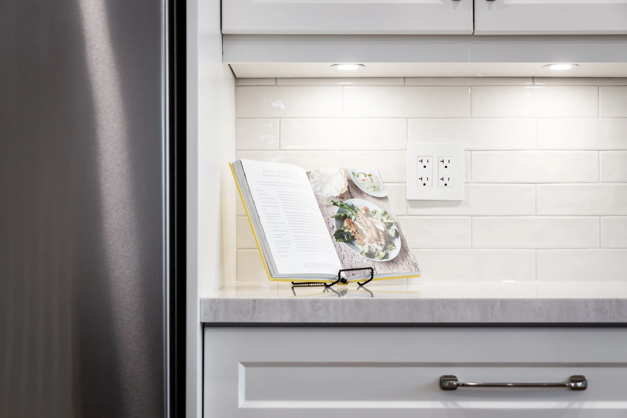 Kitchen counter detail with a cookbook stand, white subway tile, and a stainless steel refrigerator
