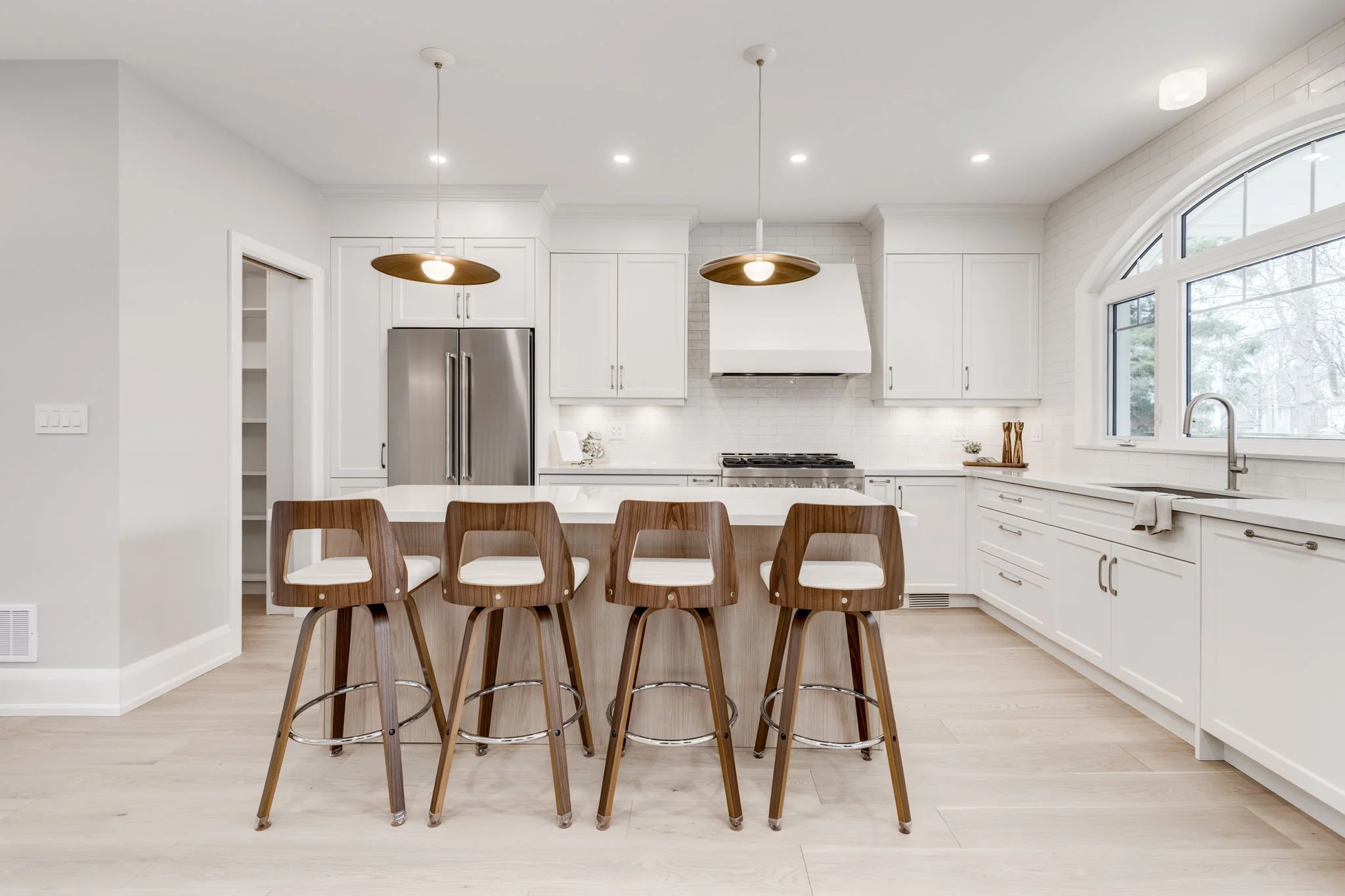 Open white kitchen with four wood bar stools at the island, brass pendants, and an arched window