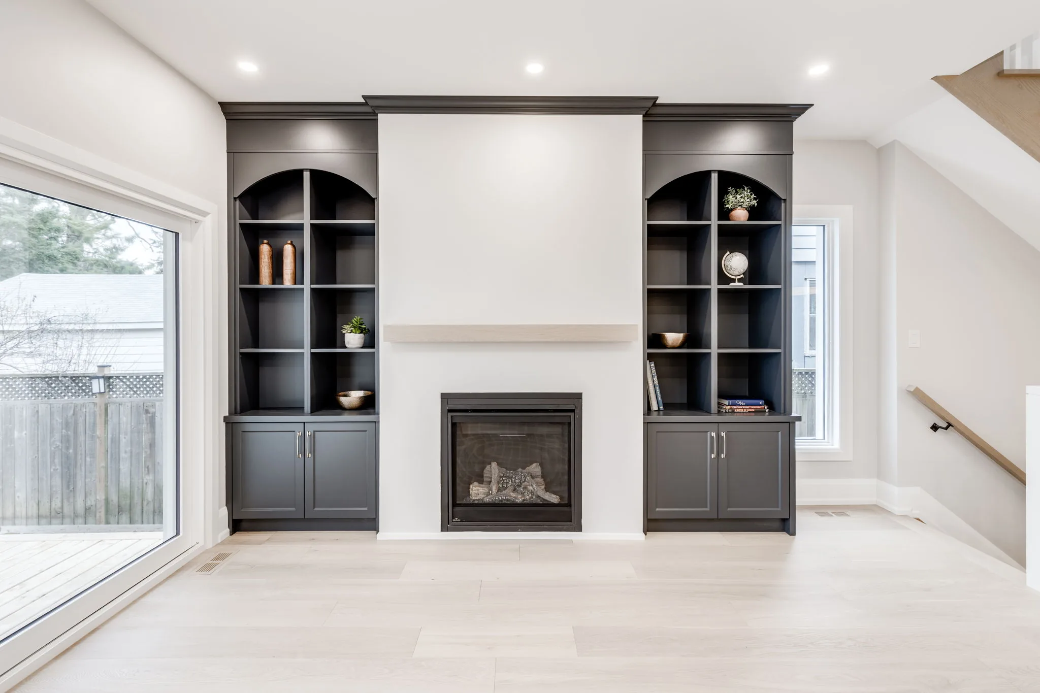 Living room after a whole-home renovation, with a gas fireplace flanked by charcoal arched built-ins, a white oak mantel, sliding doors to the deck, and the staircase beyond