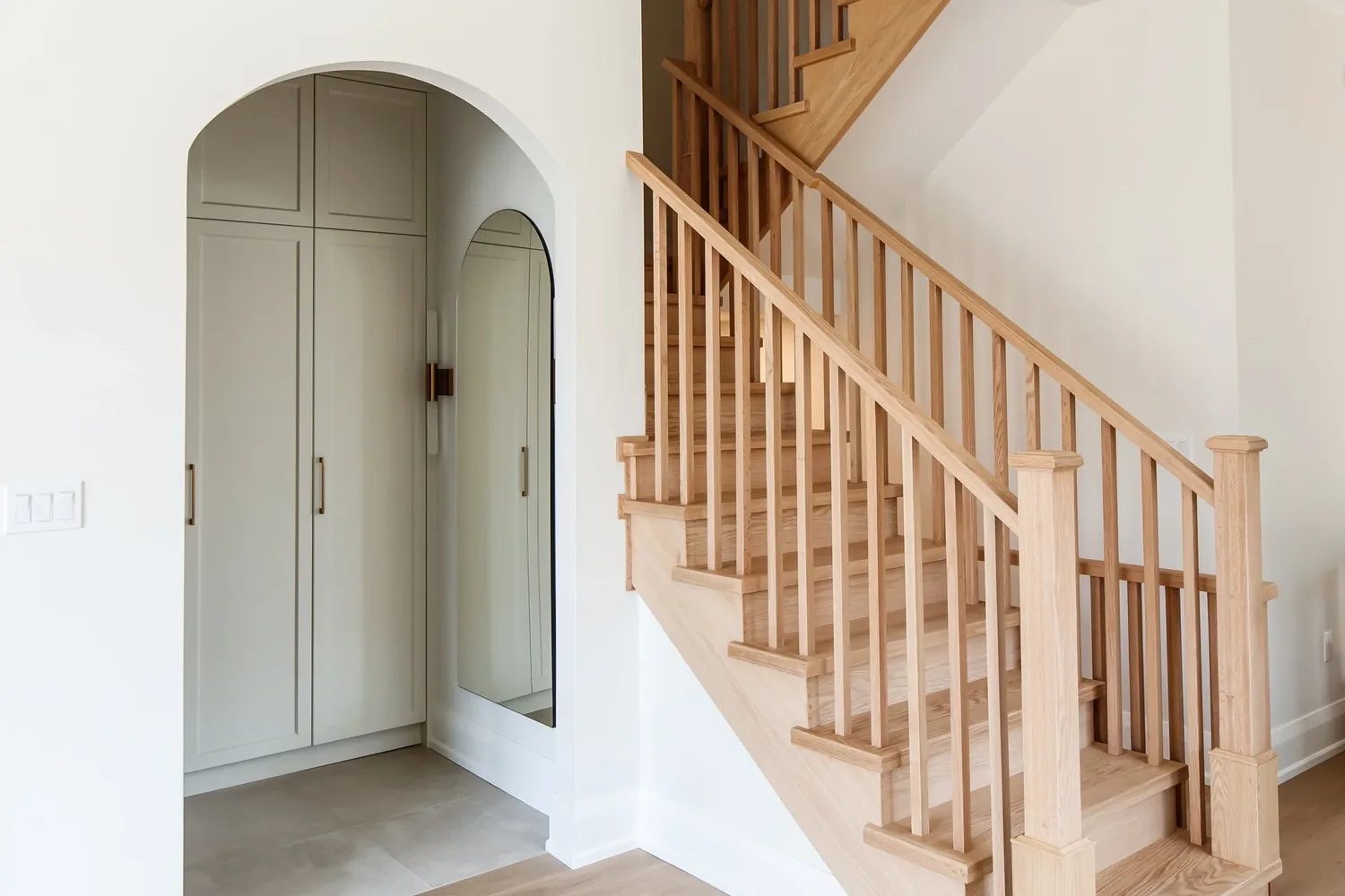 Arched doorway framing built-in cabinetry and an arched mirror beside a white oak staircase.