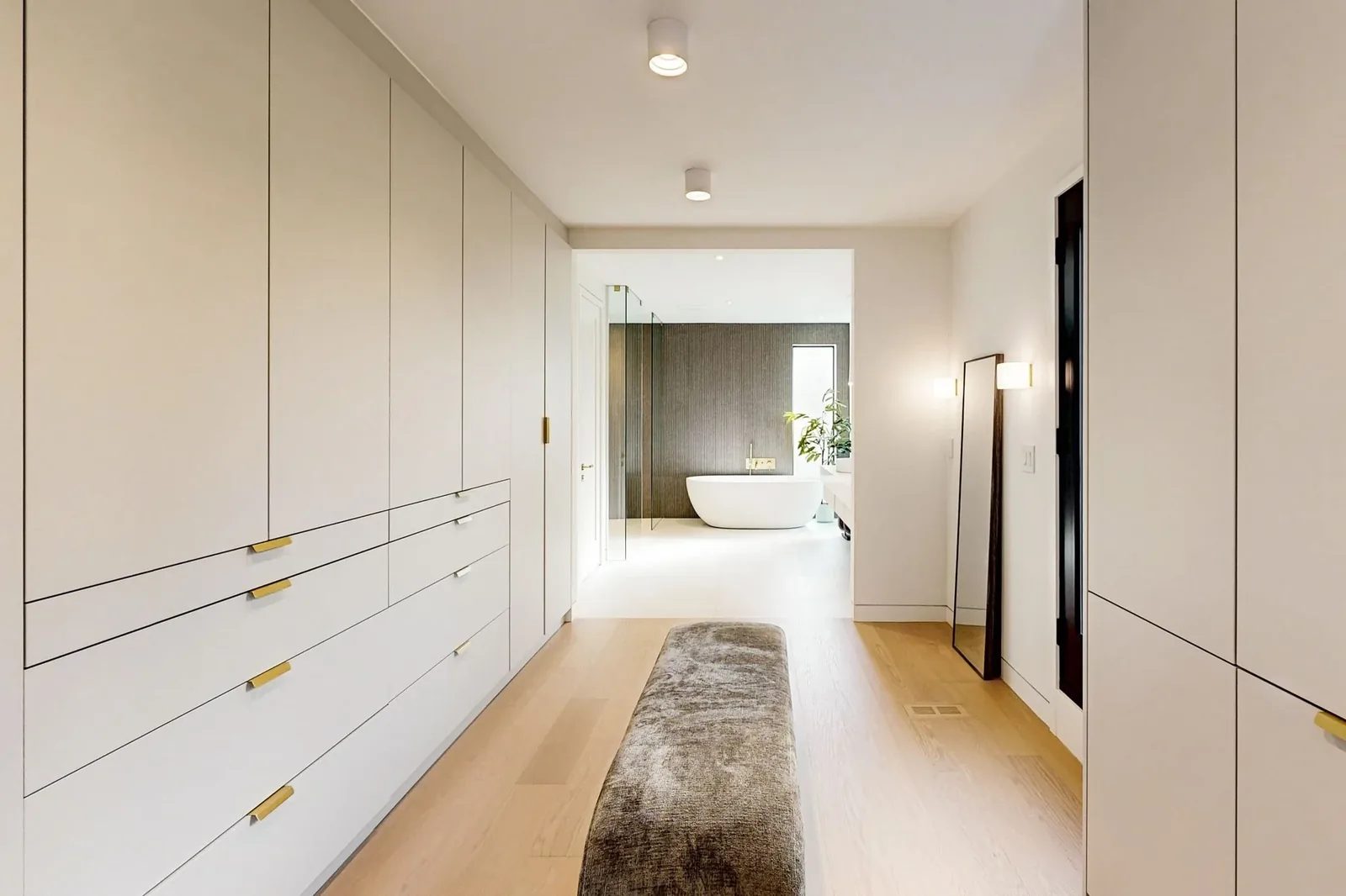 Dressing corridor with floor-to-ceiling cream wardrobes and brass pulls leading to an ensuite with a freestanding tub.