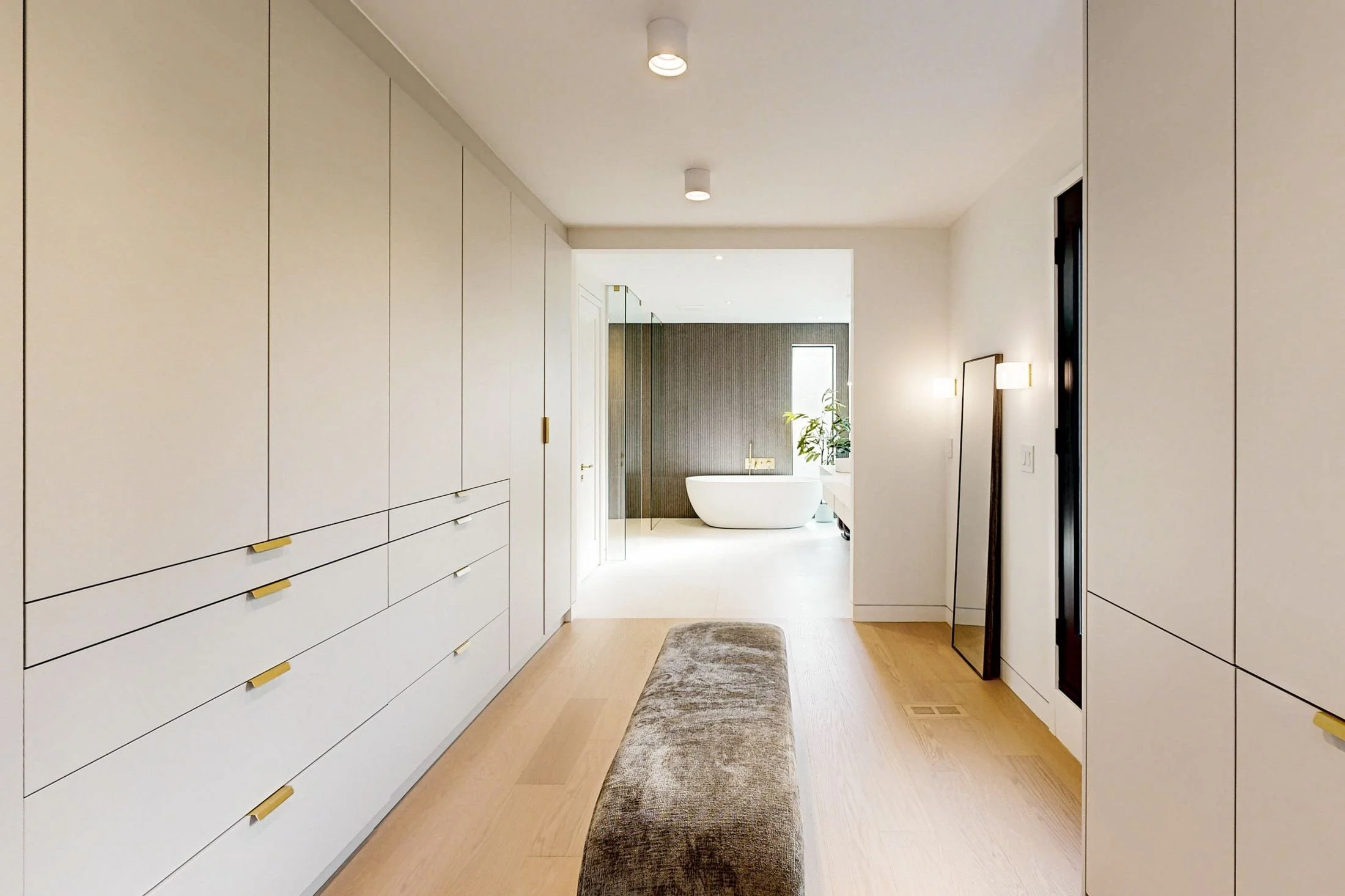 Dressing corridor with floor-to-ceiling cream wardrobes and brass pulls leading to an ensuite with a freestanding tub.