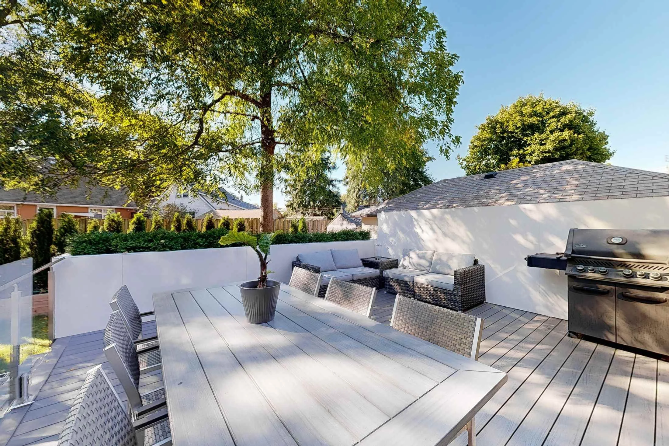Backyard deck with a grey outdoor sectional, a planter, and a stainless barbecue under a mature tree.