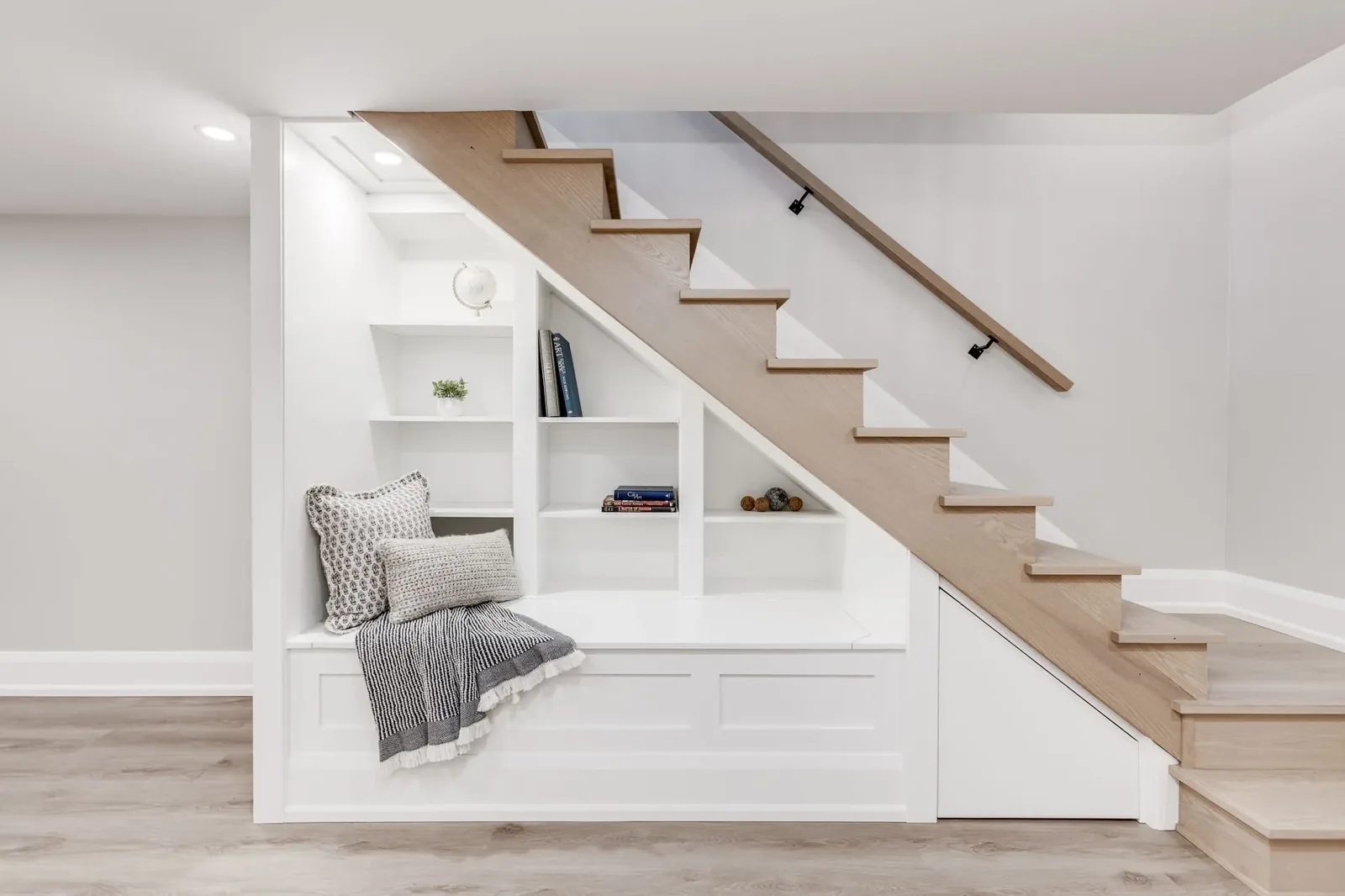 Oak and white staircase over a built-in reading nook with shelving and cushions.