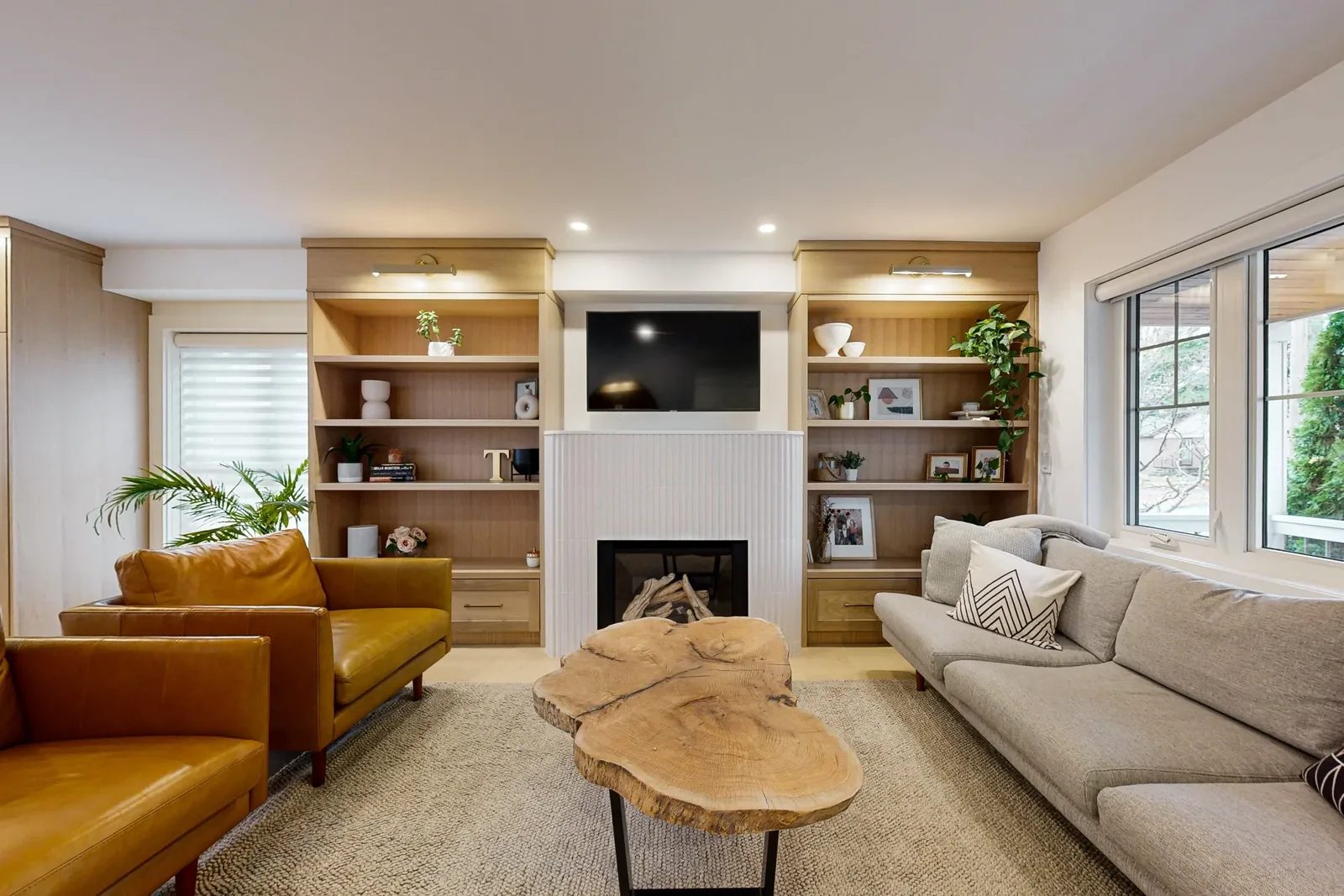 Warm living room with light-oak built-in shelving flanking a white fluted fireplace, tan leather chairs, a live-edge wood coffee table, and a soft grey sofa.