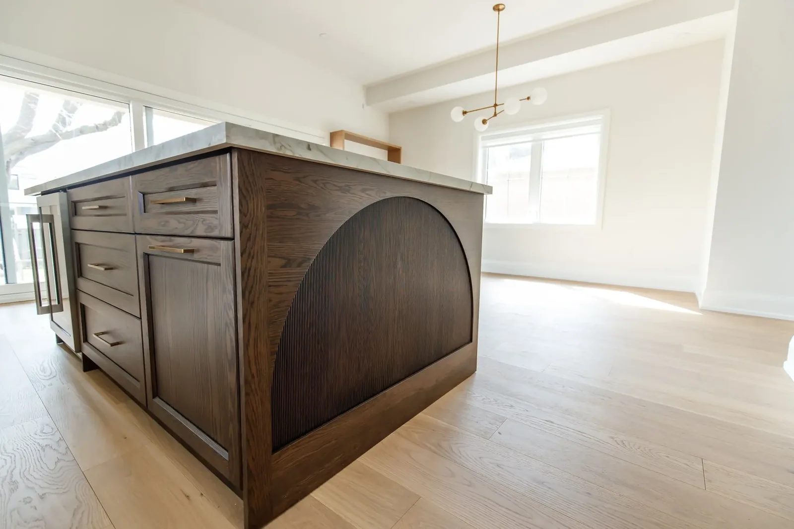 Kitchen island with an arched dark oak end panel and marble top, with a brass chandelier and wide-plank oak floors in the open dining area.