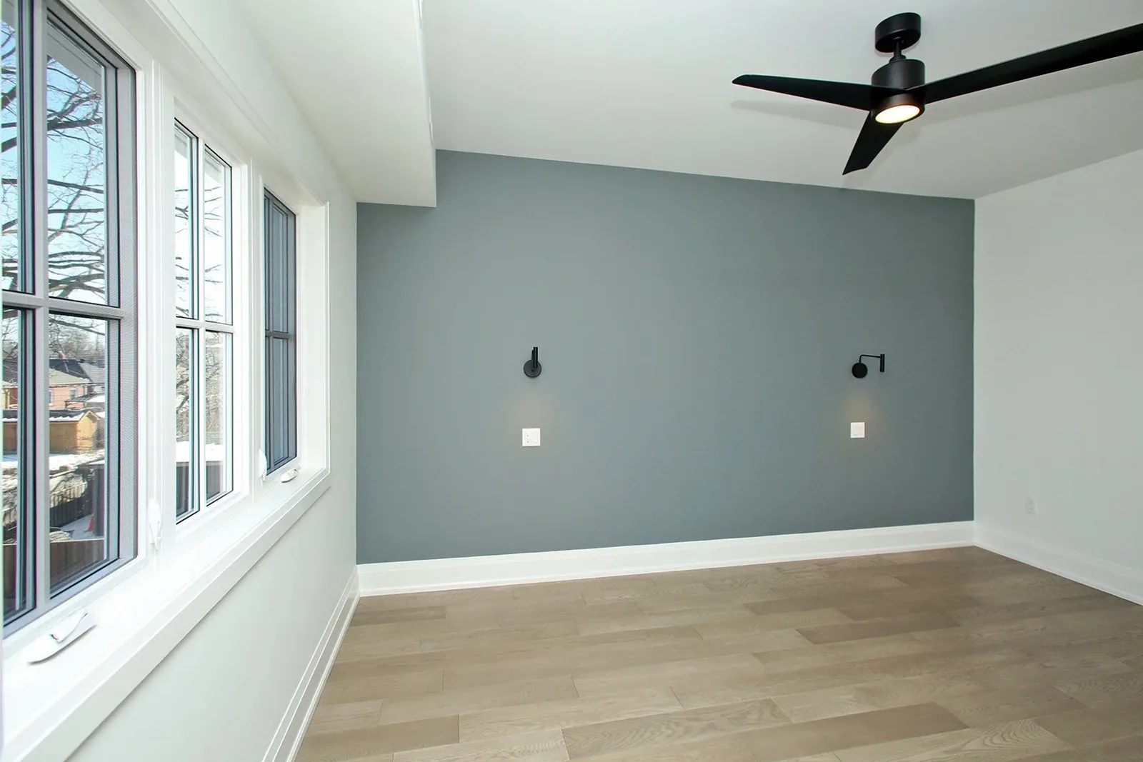 Primary bedroom with slate blue accent wall, black ceiling fan, paired sconces, and light oak hardwood.