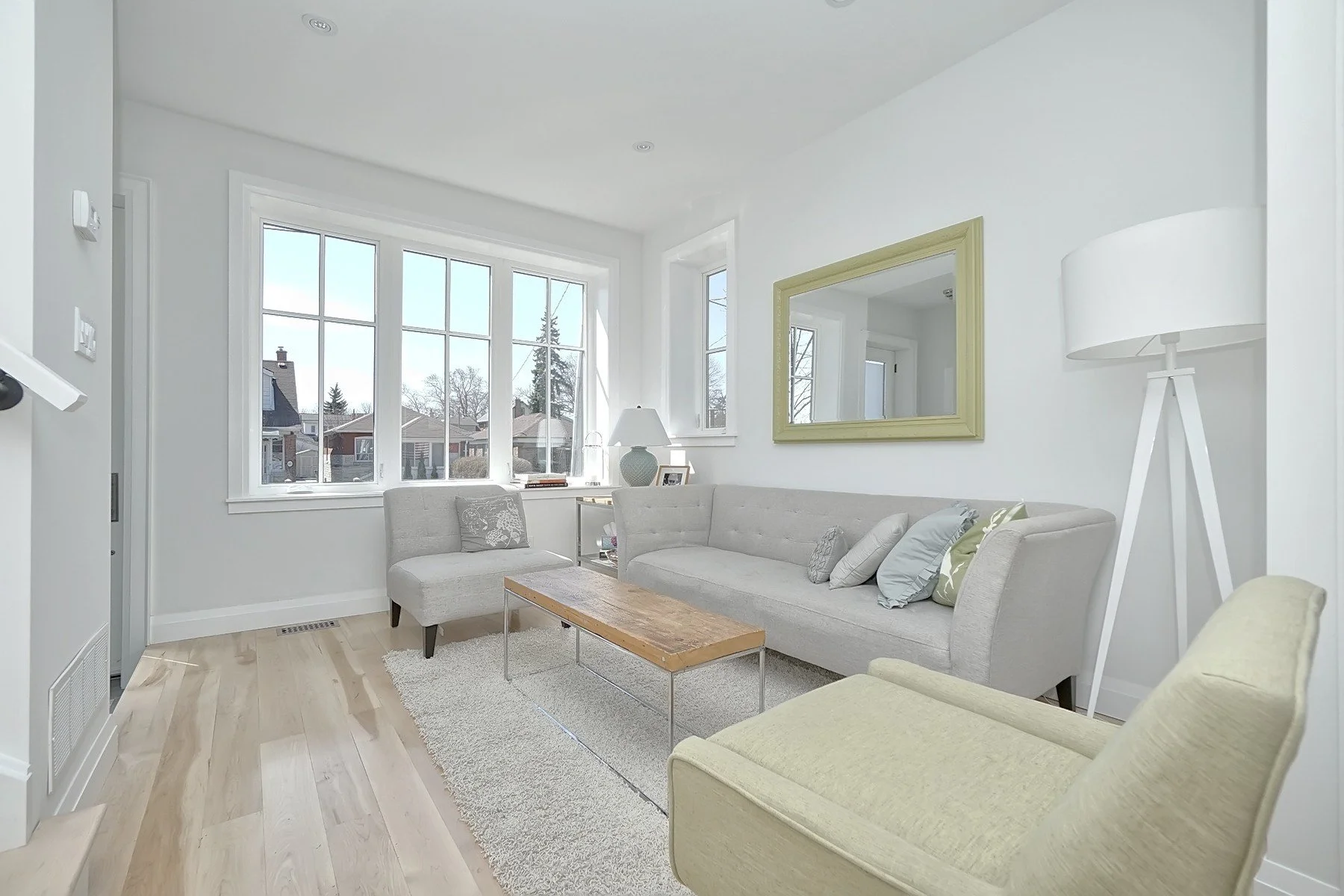 Bright Toronto living room with pale hardwood floors, large front windows, grey seating and warm wood accents.