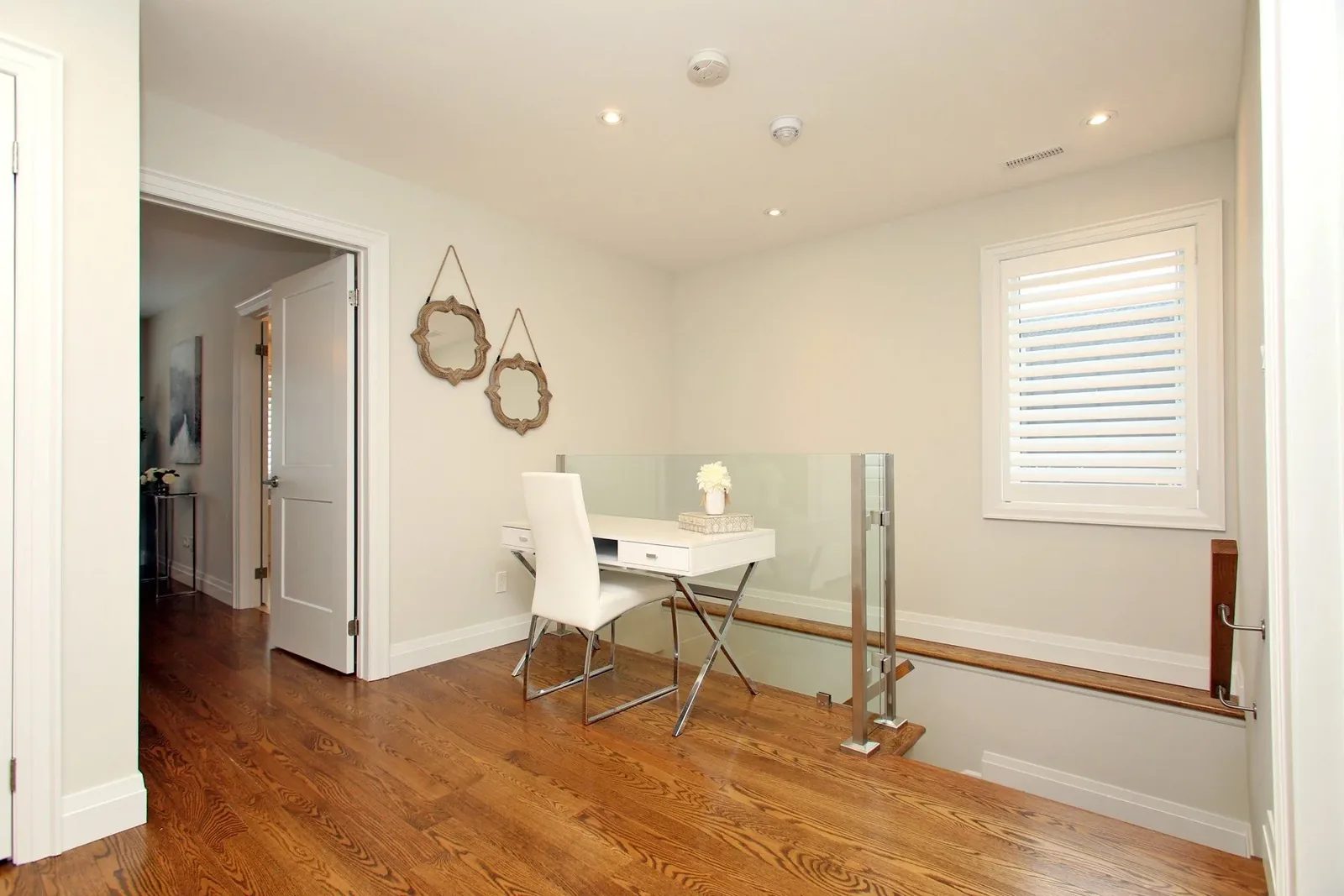 Bright upper hallway with hardwood floors, a glass stair railing, and a compact desk against warm walls.