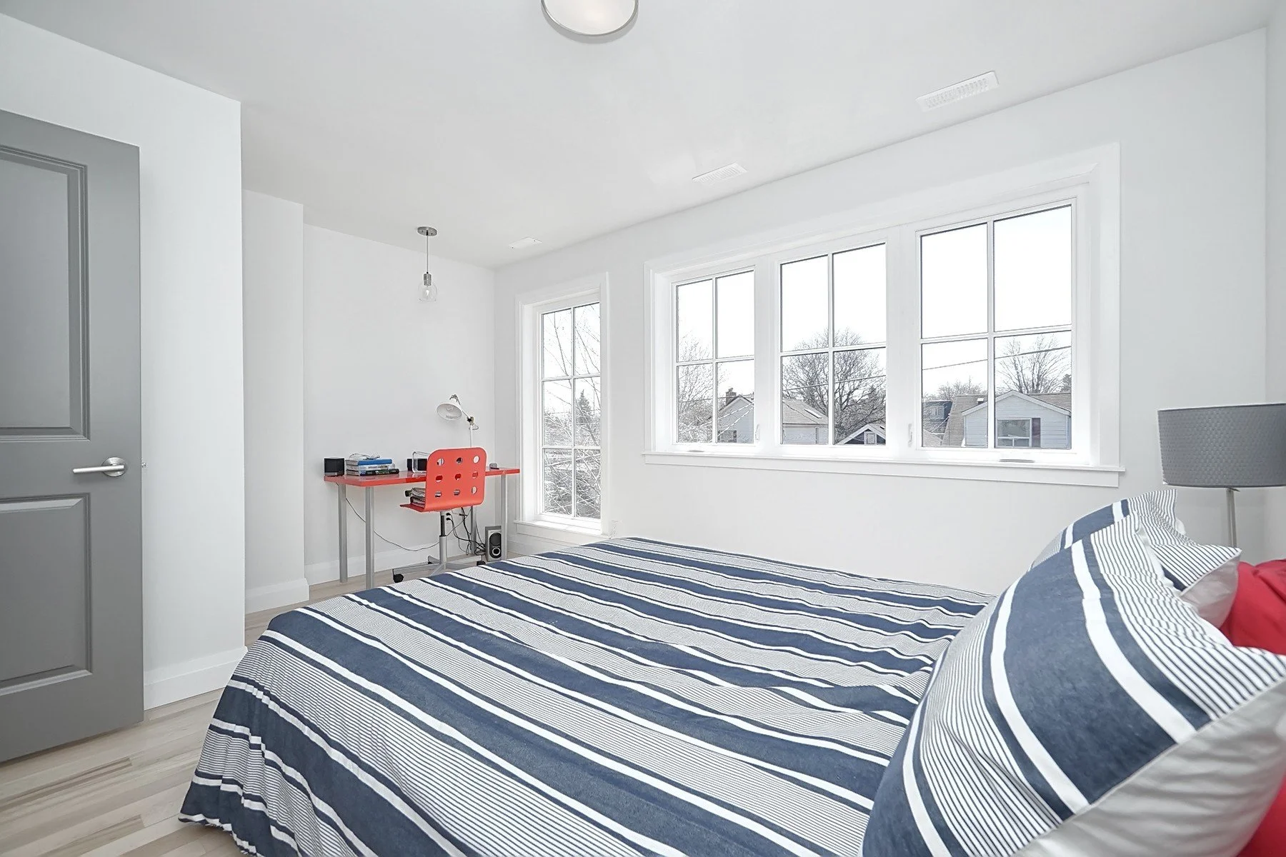 Bright white bedroom with large windows, grey door, light wood floor, and navy striped bedding.