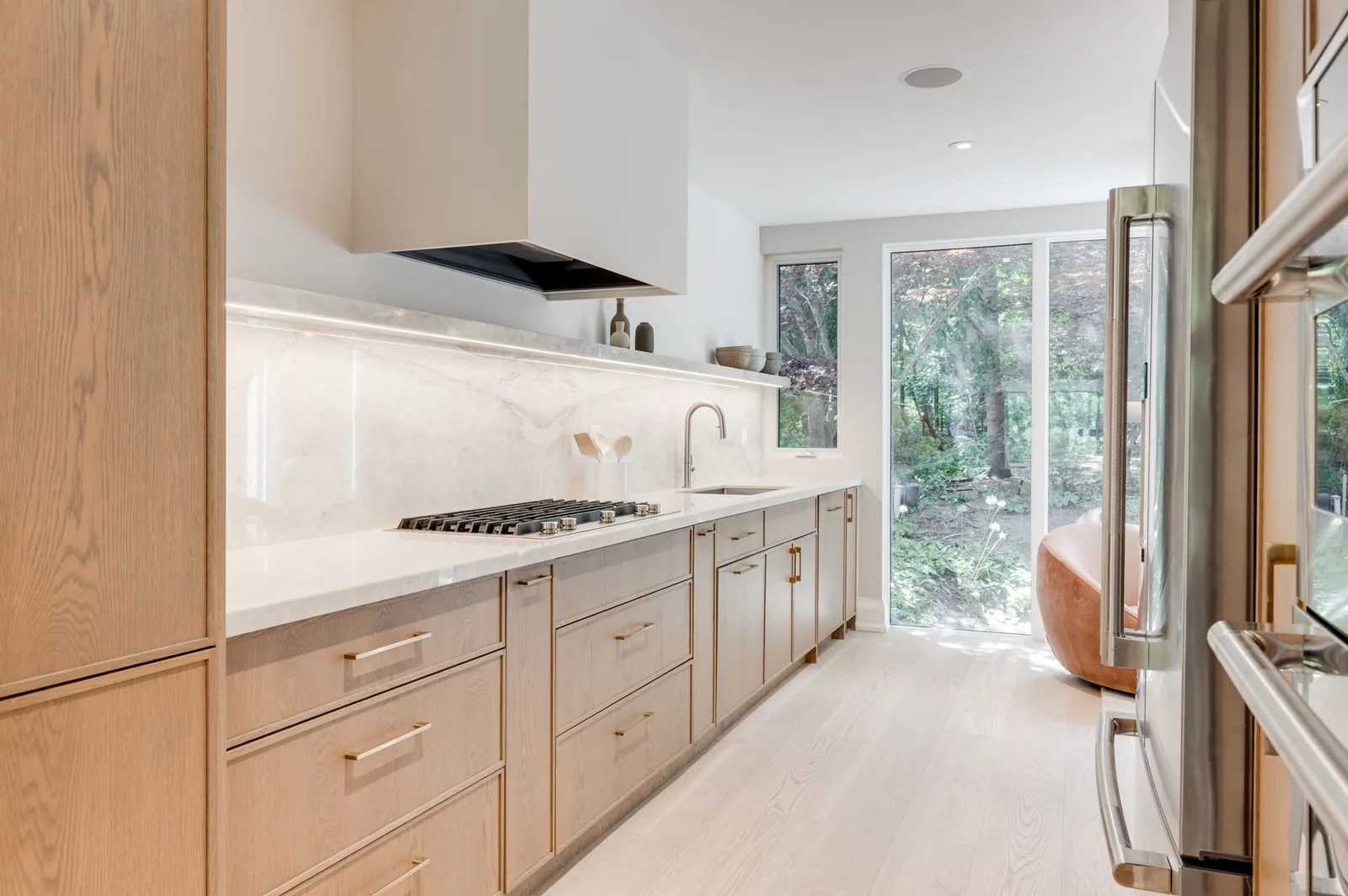 White oak galley kitchen with a marble slab backsplash, gas cooktop, and full-height glass to a wooded garden in a renovated Swansea home beside High Park.