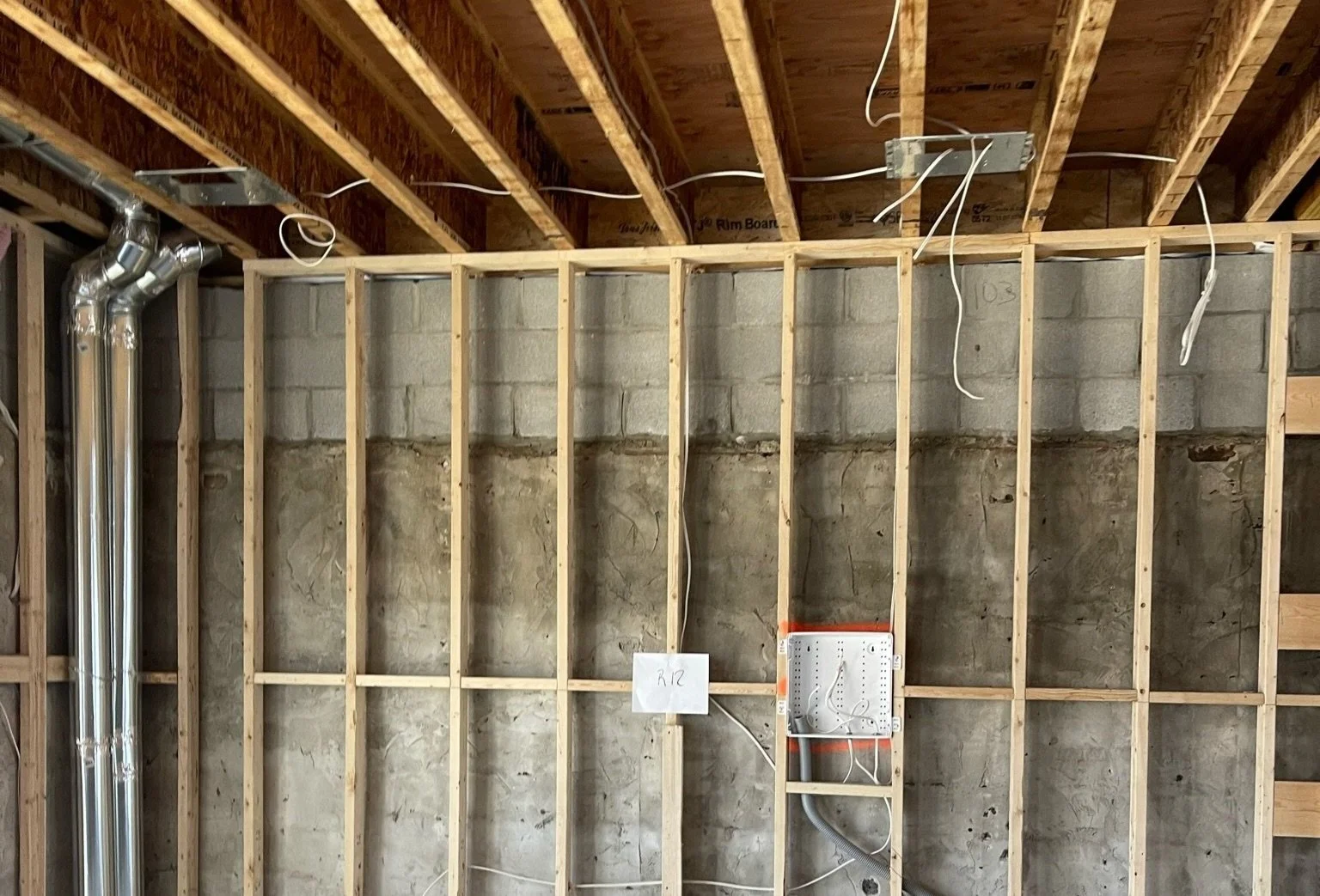 Partially constructed interior wall with exposed wood framing, electrical wiring, and metal ventilation pipes, against a concrete block wall.