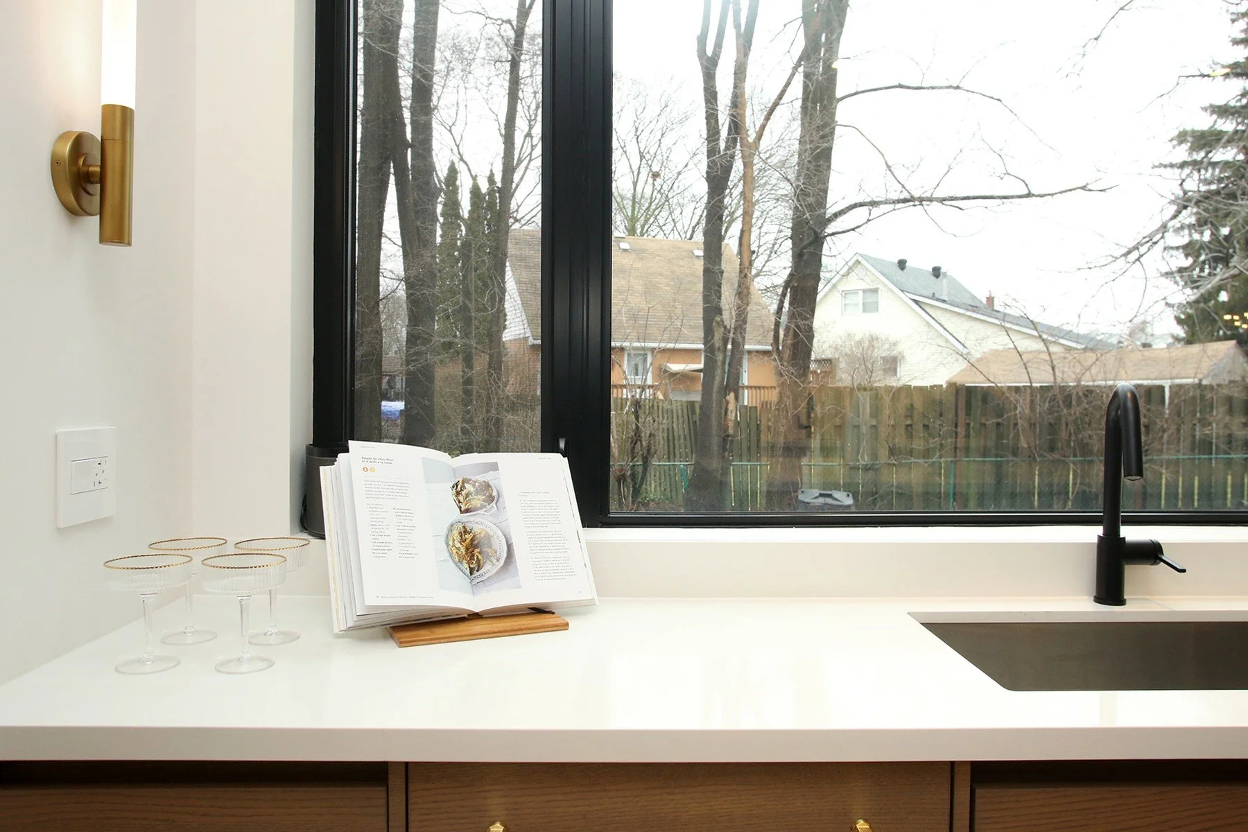 Undermount stainless sink and matte black pull-down faucet sit below a black-framed picture window with a brass sconce and coupe glasses styled on the counter.
