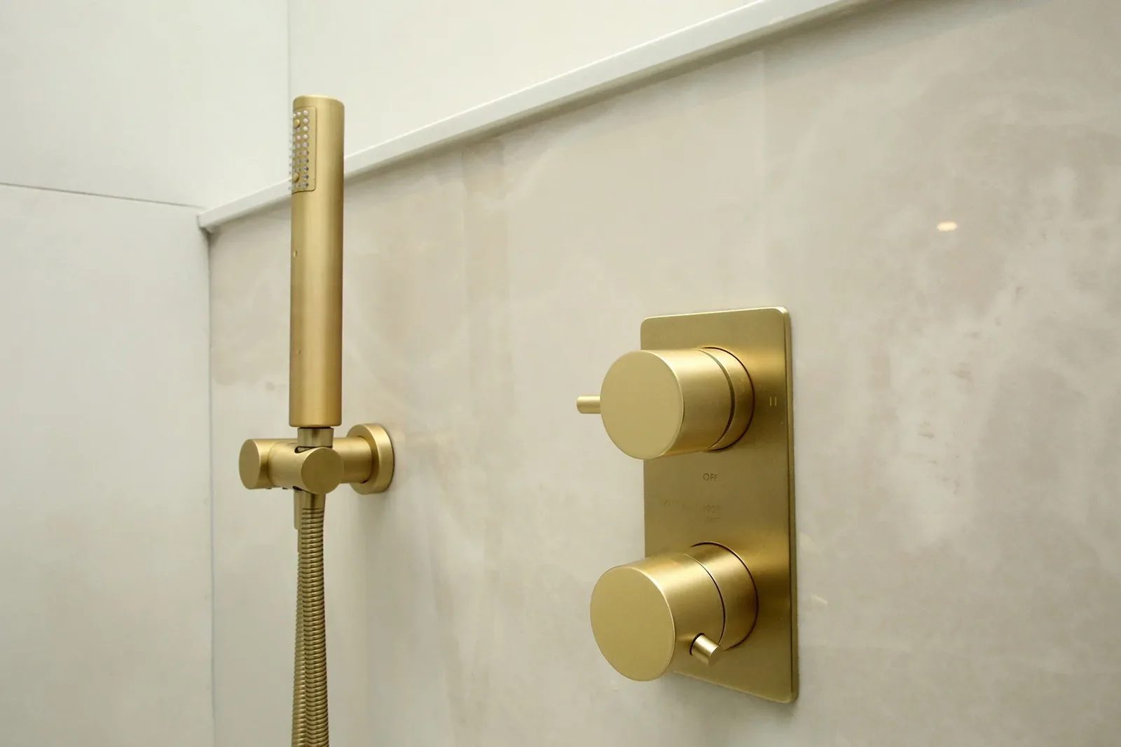 Brushed brass handheld shower and thermostatic valve on cream porcelain shower wall.