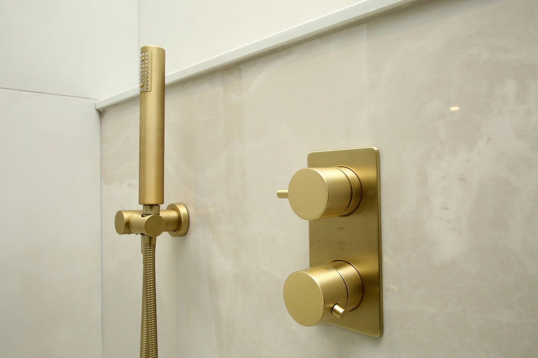 Brushed brass handheld shower and thermostatic valve on cream porcelain shower wall.