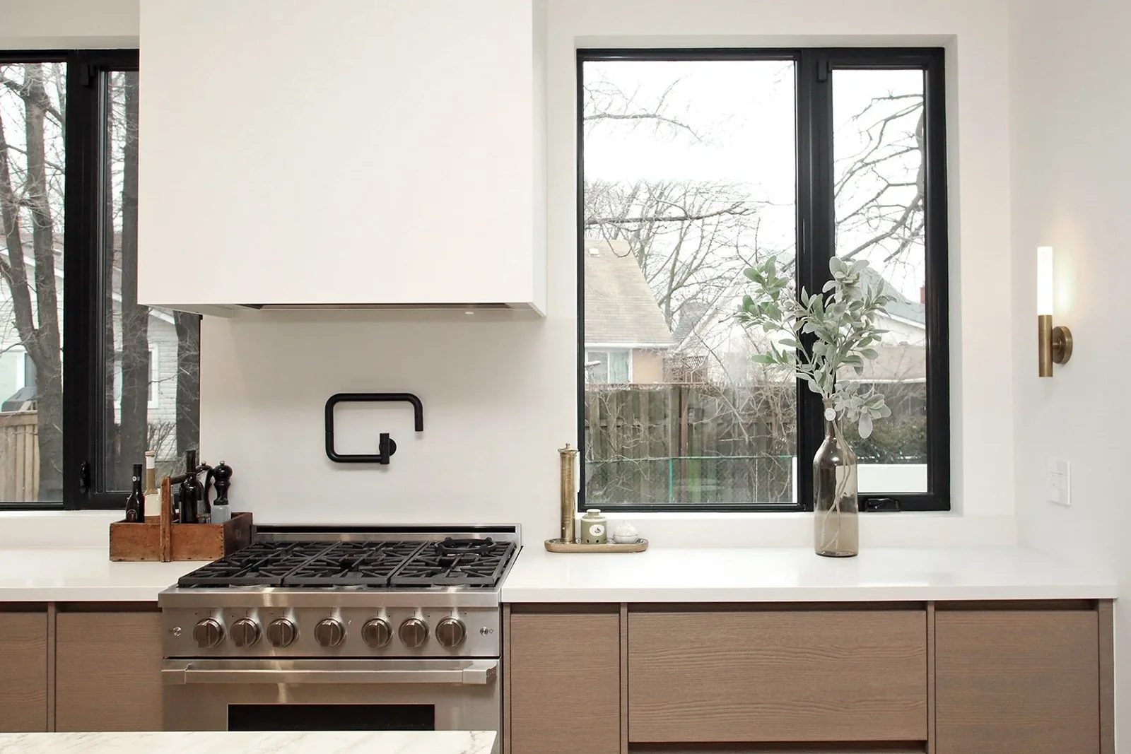Symmetrical view of a stainless gas range flanked by oak cabinetry under a bright picture window with a matte black pot filler.
