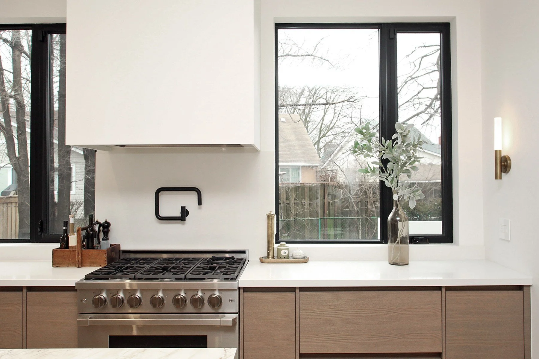 Symmetrical view of a stainless gas range flanked by oak cabinetry under a bright picture window with a matte black pot filler.