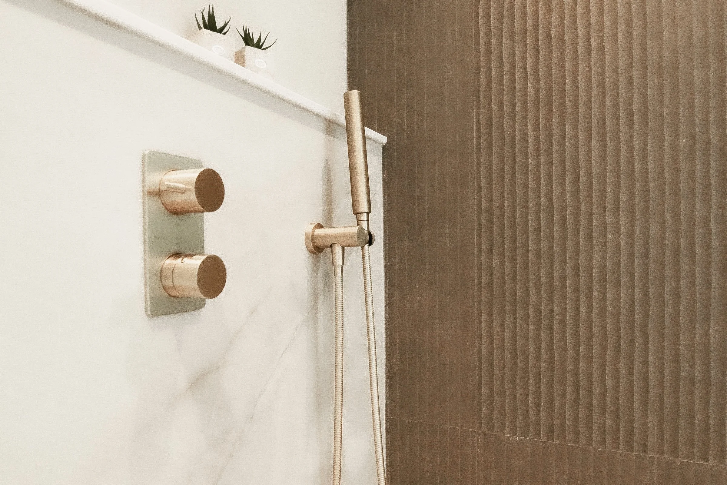 Close-up of a brushed brass thermostatic valve and handheld sprayer set against veined marble-look stone with a fluted taupe tile wall behind.
