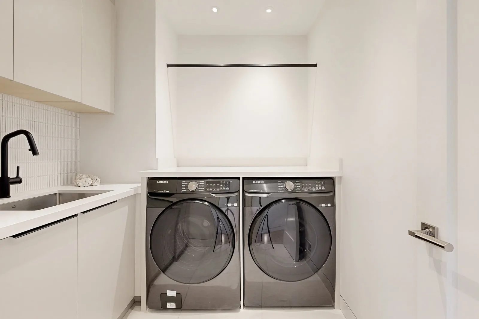 Modern laundry room with a Samsung washer and dryer pair under a quartz counter a matte black faucet over a stainless sink and a hanging rod above.
