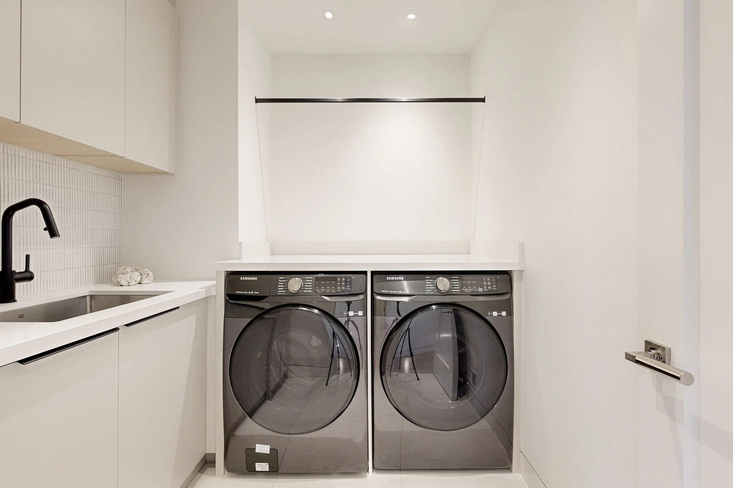 Modern laundry room with a Samsung washer and dryer pair under a quartz counter a matte black faucet over a stainless sink and a hanging rod above.