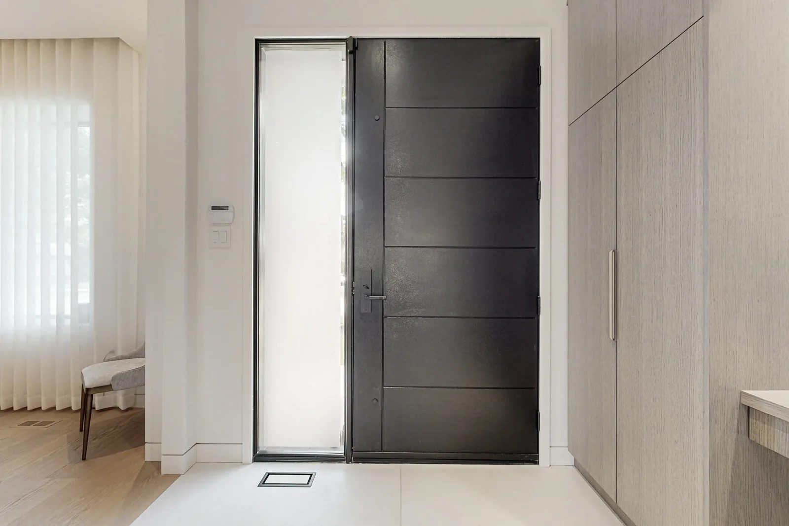 Interior view of a tall matte black pivot entry door beside a built in oak veneer closet wall.