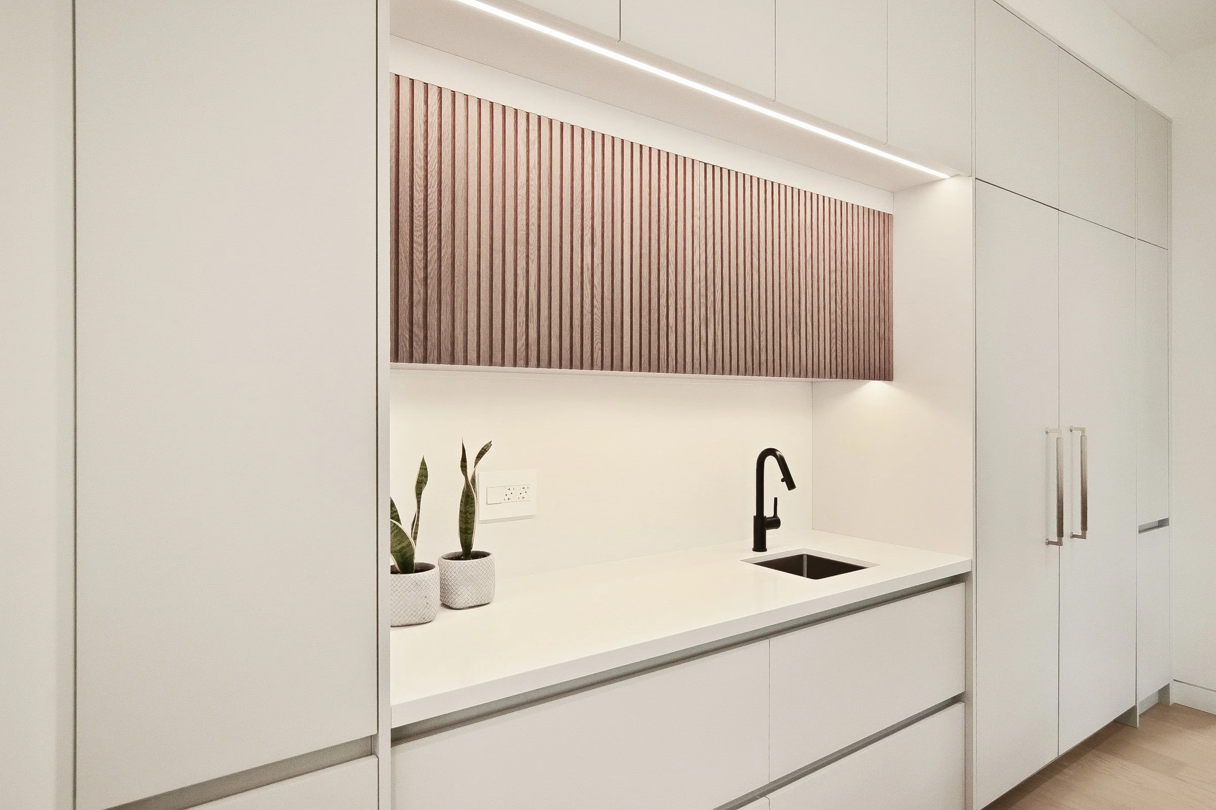 Minimalist servery nook with fluted walnut panel matte black faucet and handleless white cabinetry.