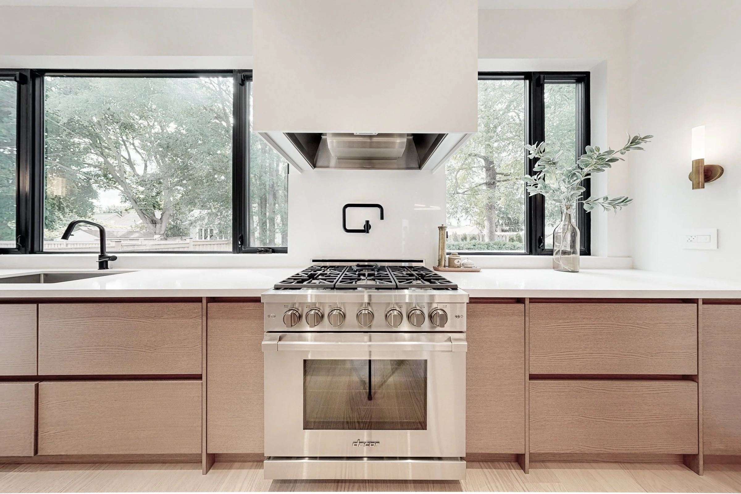 Professional stainless gas range framed by light oak cabinetry and black framed picture windows.