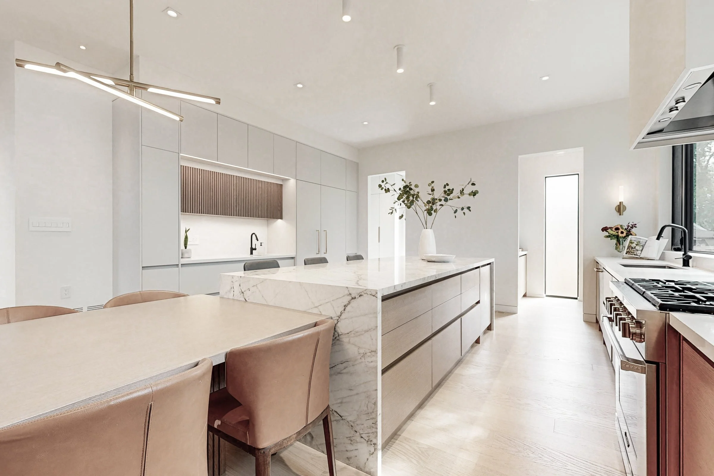 Open concept kitchen and dining with oversized marble waterfall island light oak cabinetry and linear chandelier.