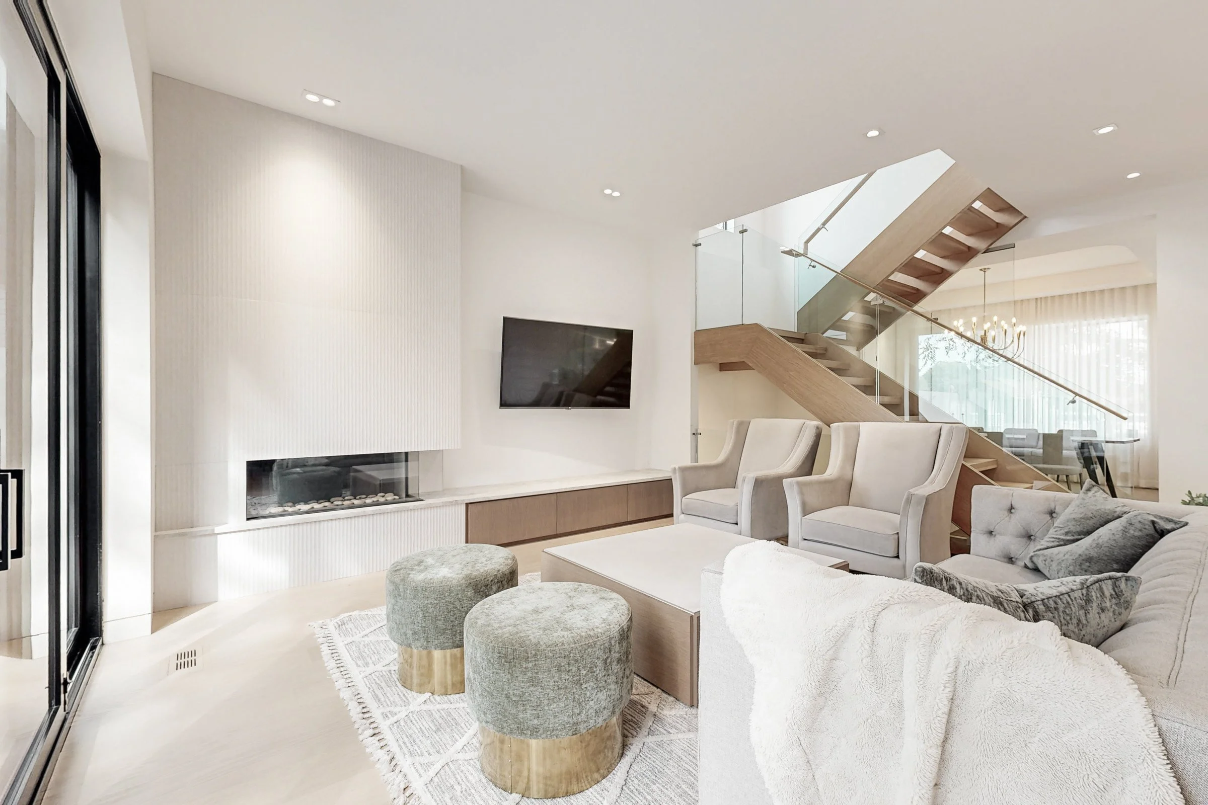 Living room with tufted sofa, velvet ottomans, and fluted tile fireplace beside a floating oak and glass staircase.