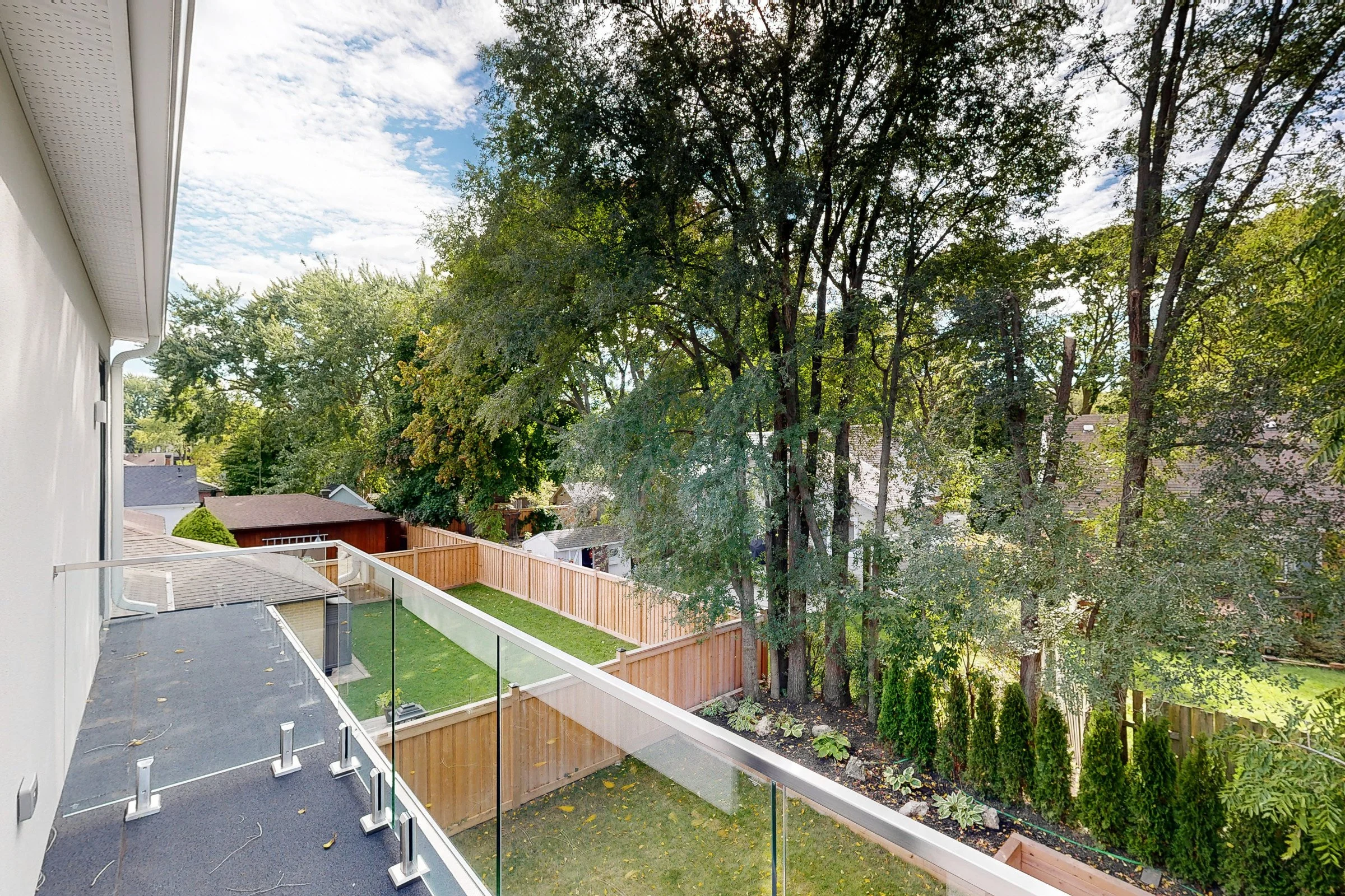 View from a second-storey balcony with frameless glass railing looking down over a landscaped backyard and cedar privacy fence.