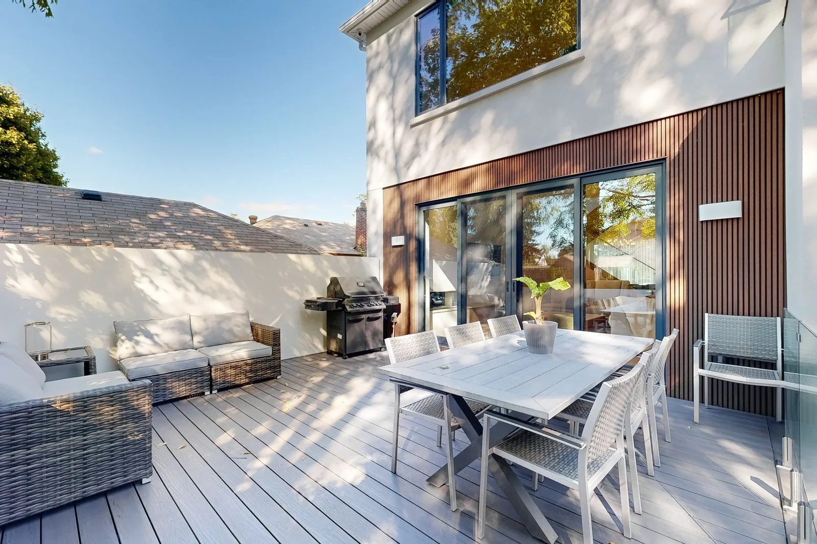 Contemporary rear elevation with white stucco cedar slat cladding and black-framed sliding doors opening onto a deck with a dining set and grill.