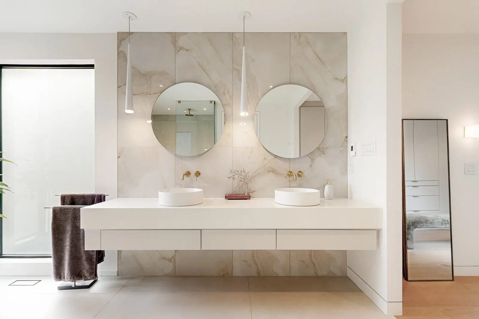 Floating matte white double vanity with twin vessel sinks brushed brass faucets and round mirrors set against a veined porcelain slab wall.