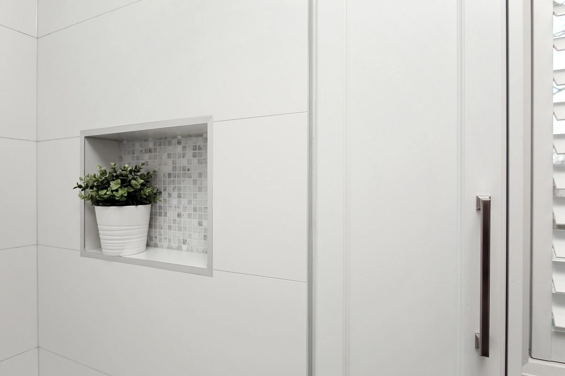 White shower surround with a recessed mosaic niche, clean glass enclosure, and a small potted plant.