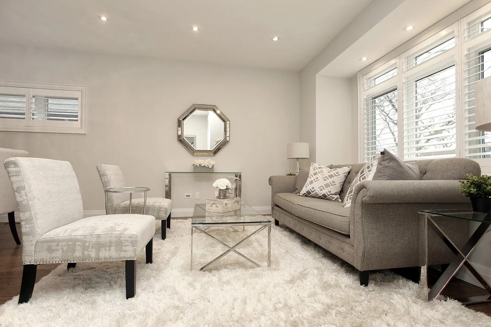 Bright neutral living room with a plush rug, glass tables, recessed lights and shuttered windows.