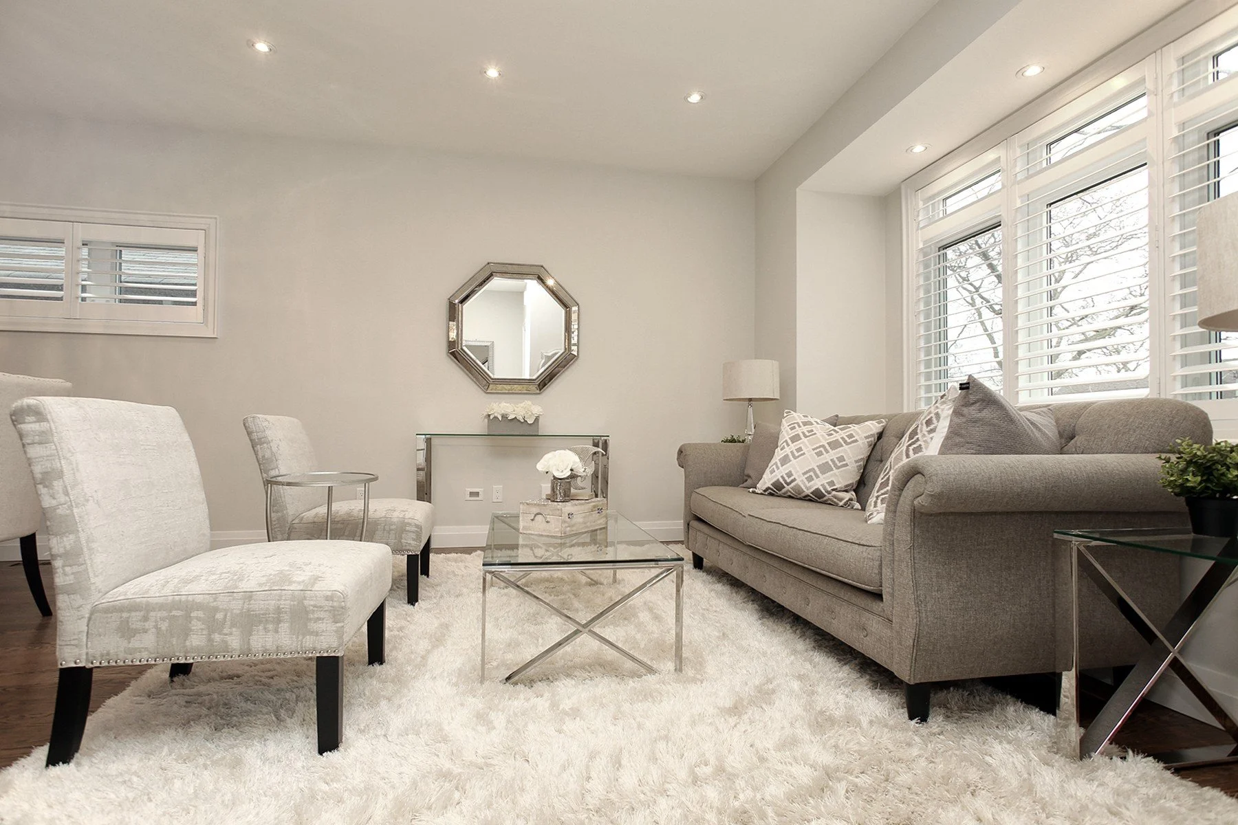 Bright neutral living room with a plush rug, glass tables, recessed lights and shuttered windows.
