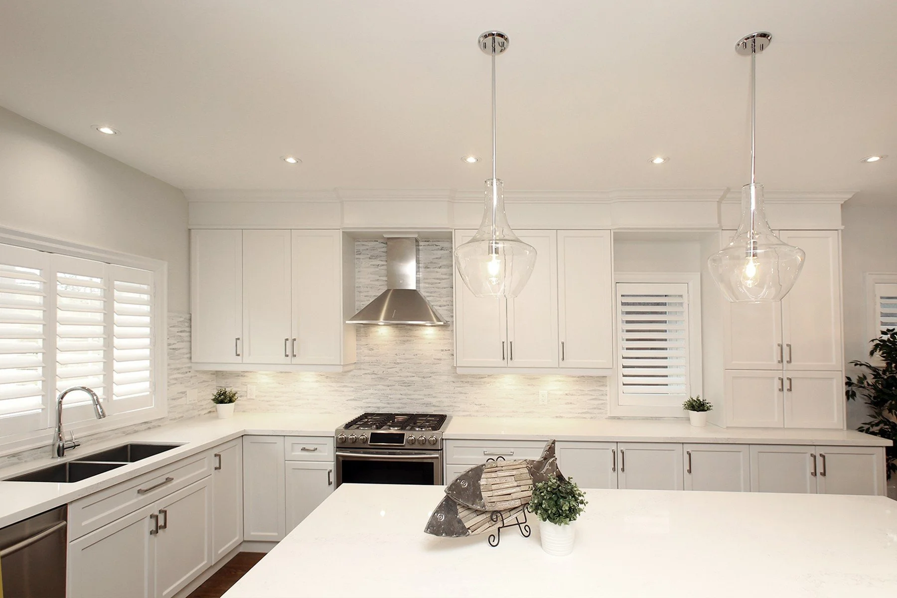 Bright white kitchen with shaker cabinets, quartz island, glass pendants and plantation shutters.