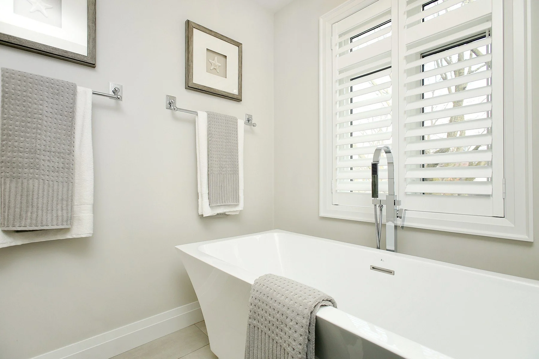 Bright bathroom with a freestanding tub, chrome filler, grey towels, and white shutters.
