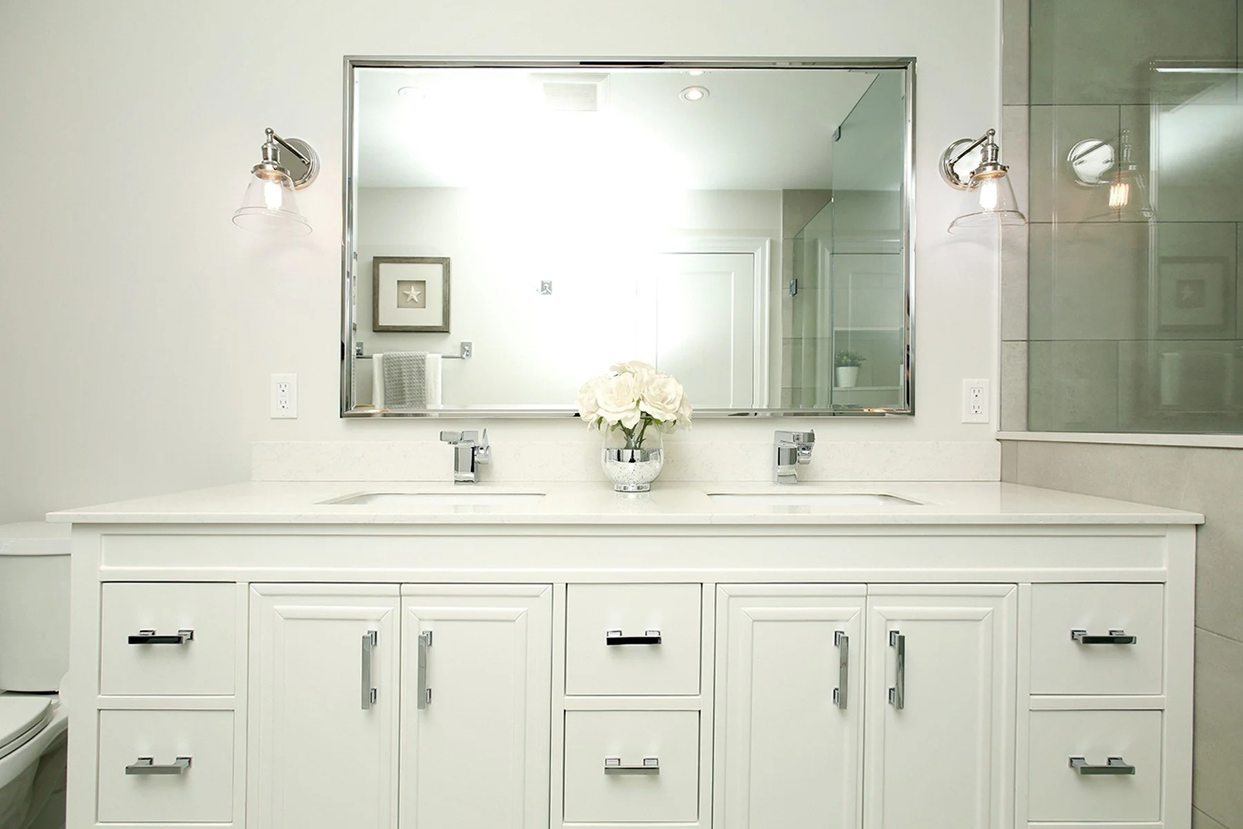 Bright white bathroom renovation with a double vanity, chrome sconces, a large mirror, and soft grey tile.