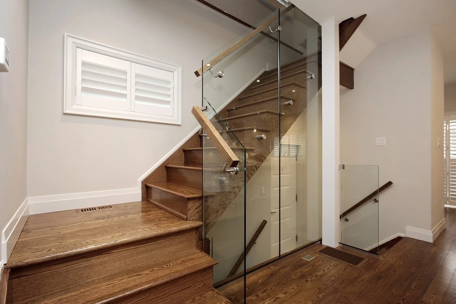 Warm white entry stair with natural oak treads, glass railings, and plantation shutters in a finished Toronto home.