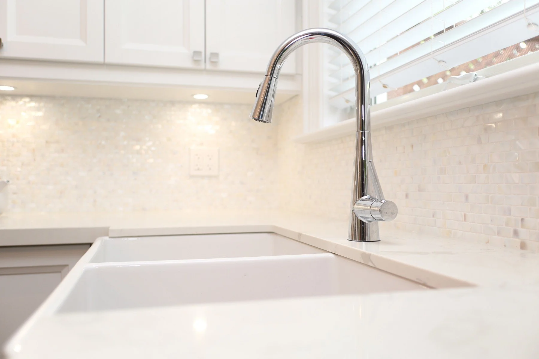 Polished chrome kitchen faucet over a white farmhouse sink with warm tile backsplash and bright window light.