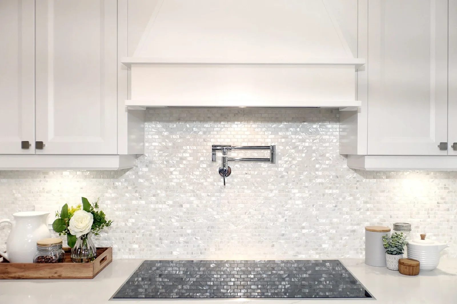White kitchen cooktop wall with iridescent tile backsplash, pot filler, shaker cabinetry and soft lighting.