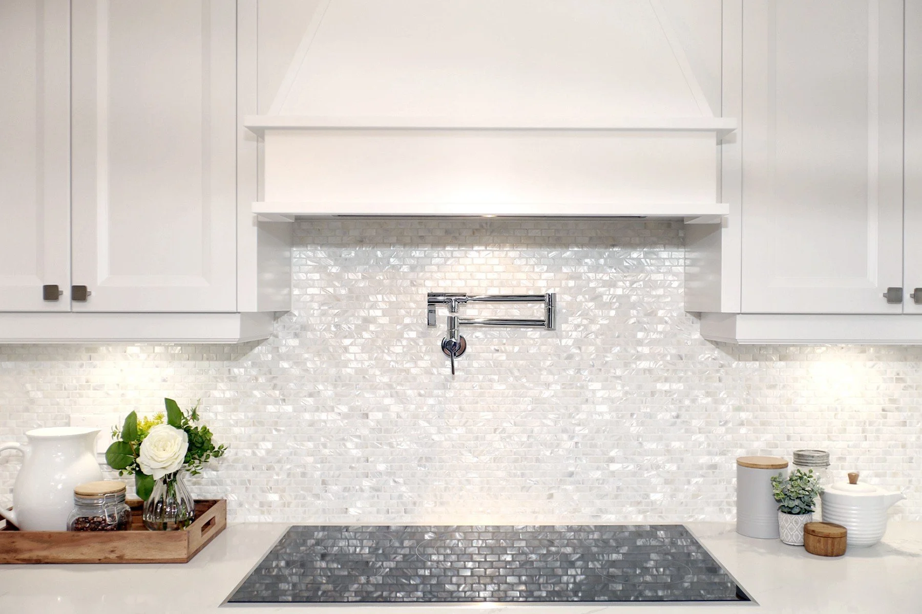 White kitchen cooktop wall with iridescent tile backsplash, pot filler, shaker cabinetry and soft lighting.
