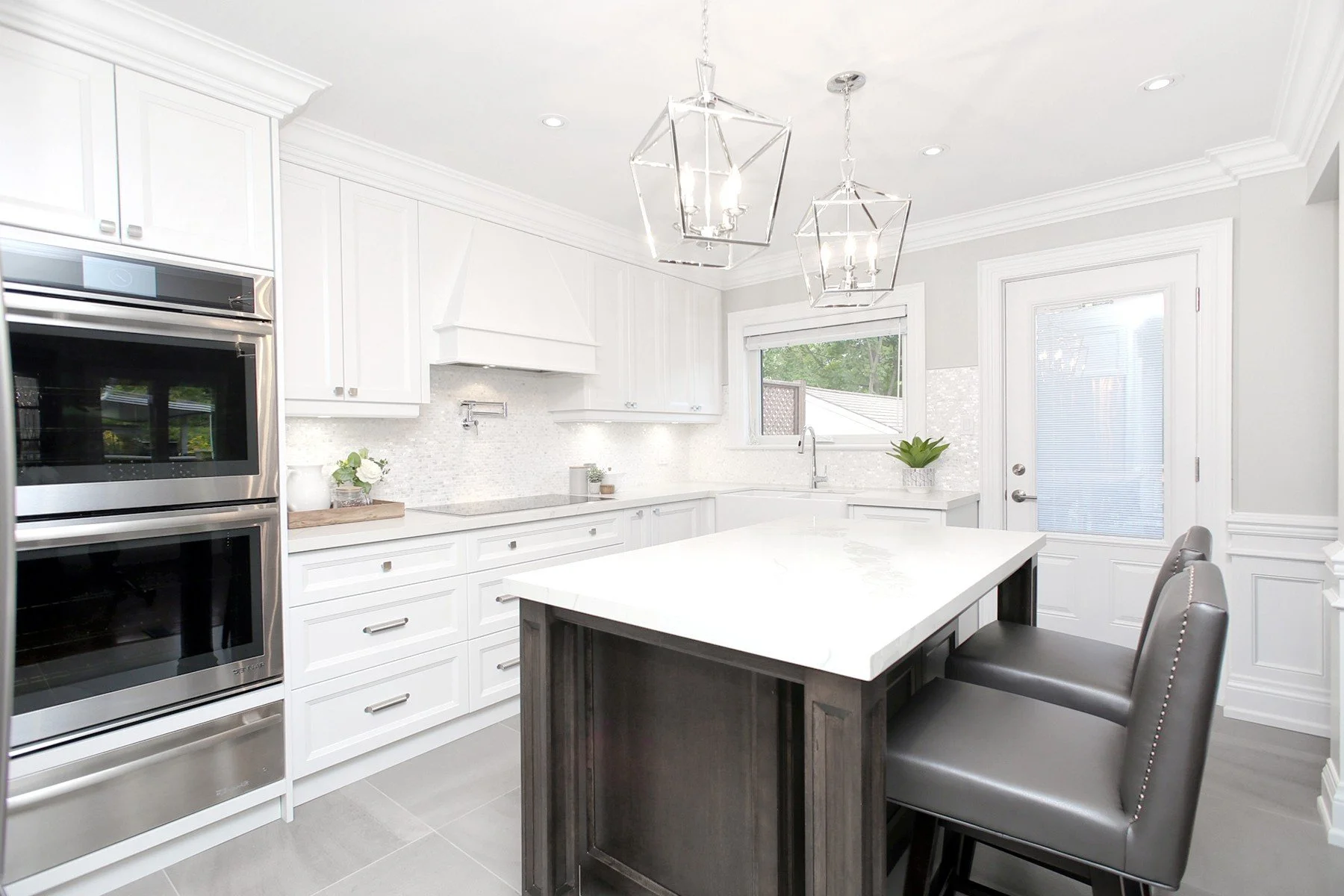 Bright white kitchen renovation with quartz island, wall ovens, grey stools and glass pendant lights.