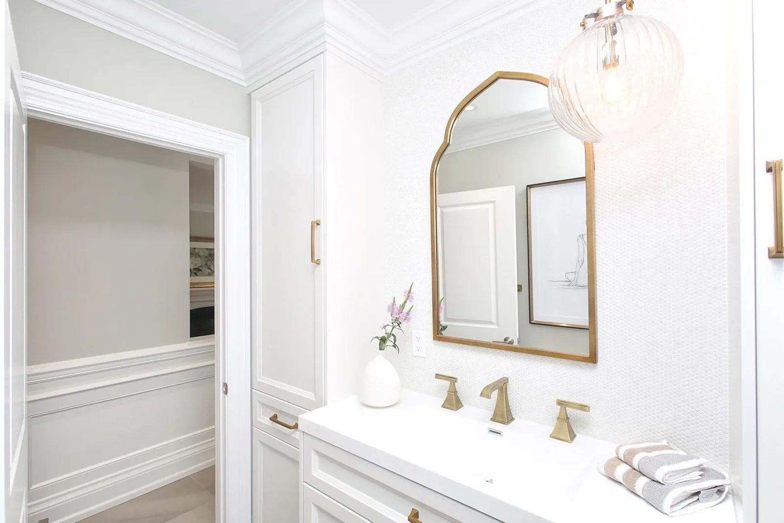 Bright finished bathroom vanity with white millwork, brass fixtures, textured walls and an arched mirror.