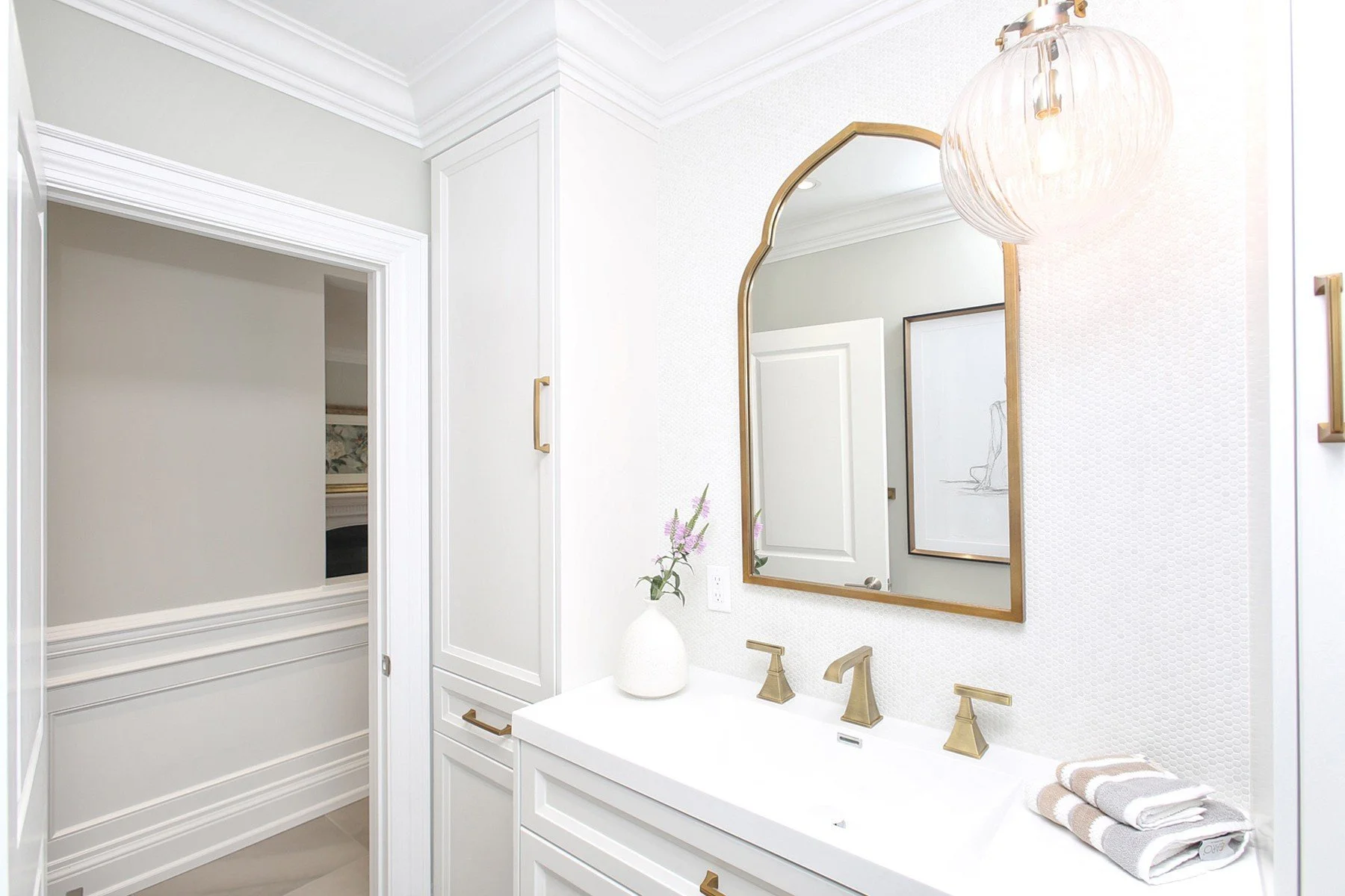 Bright finished bathroom vanity with white millwork, brass fixtures, textured walls and an arched mirror.