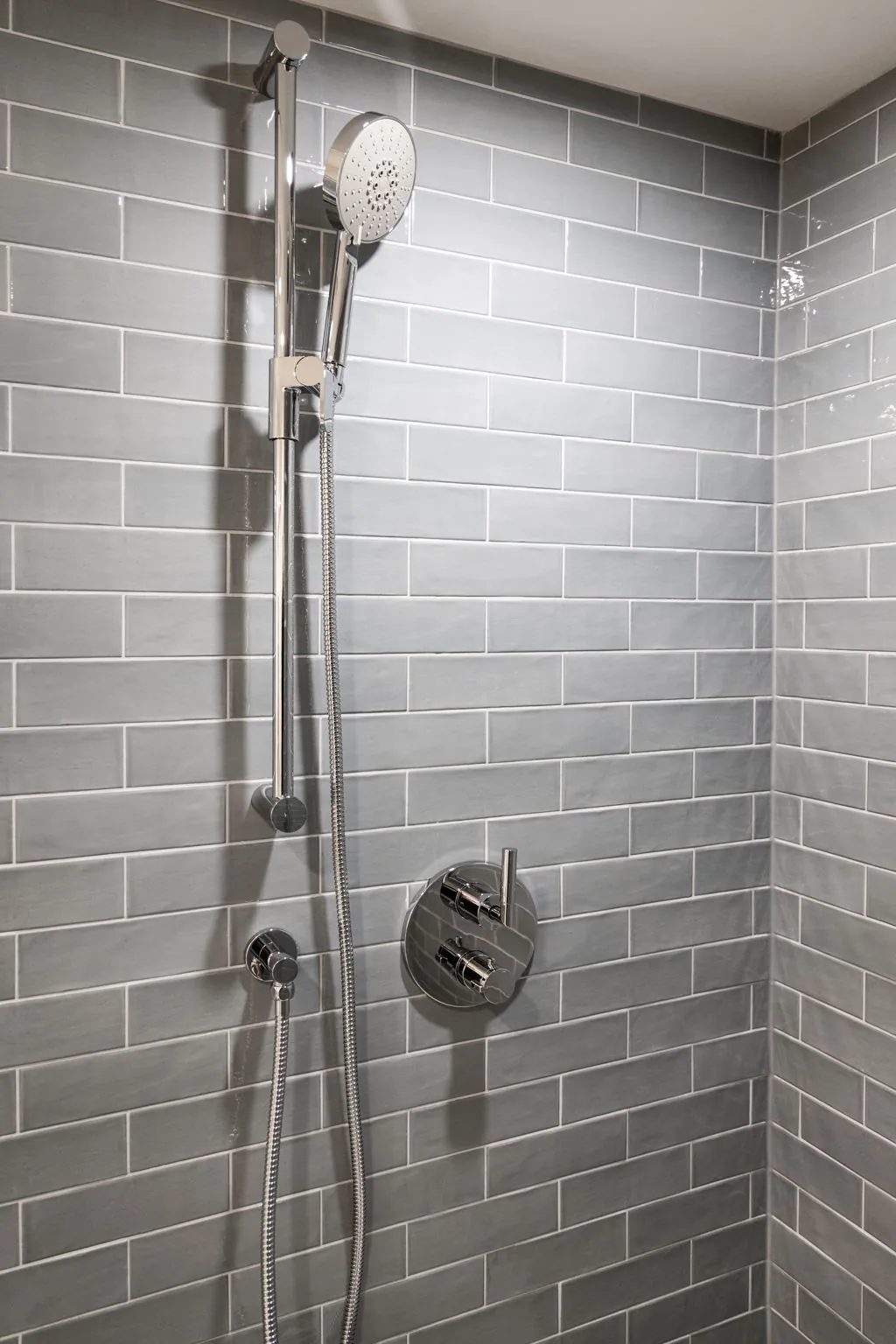 Chrome hand shower on a riser bar with a round thermostatic valve, set against blue-grey subway tile.