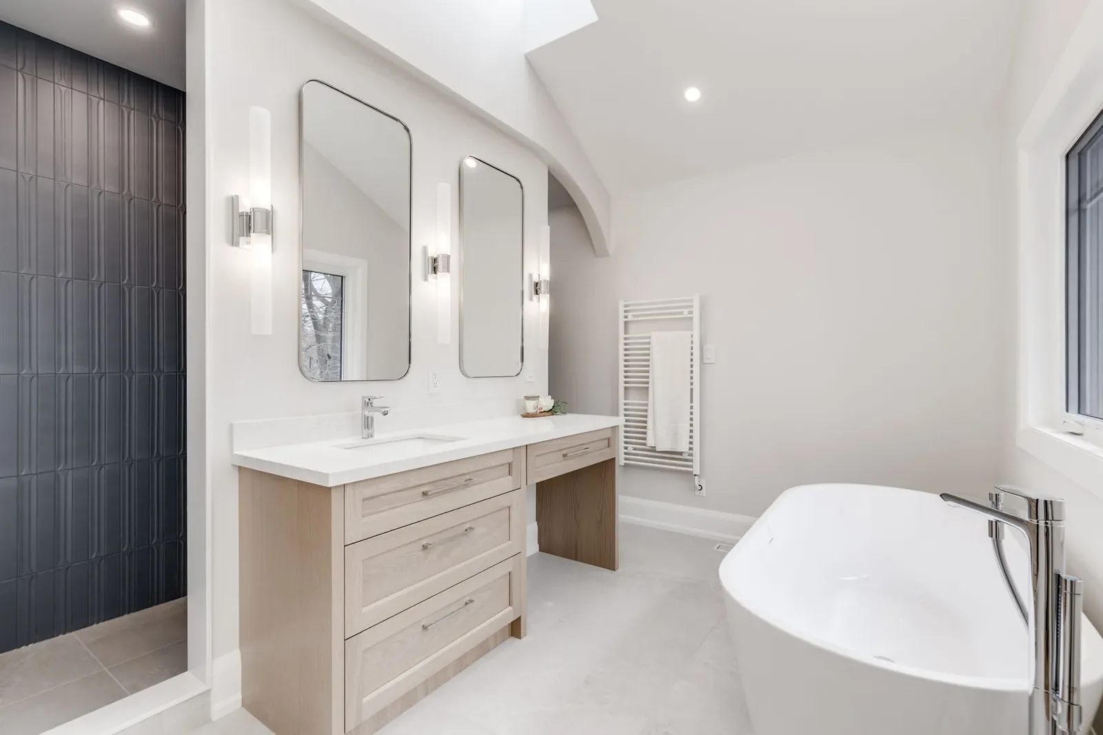 Primary ensuite with a light oak vanity and makeup counter, twin chrome-framed mirrors and sconces, the dark fluted tile shower and the freestanding tub.